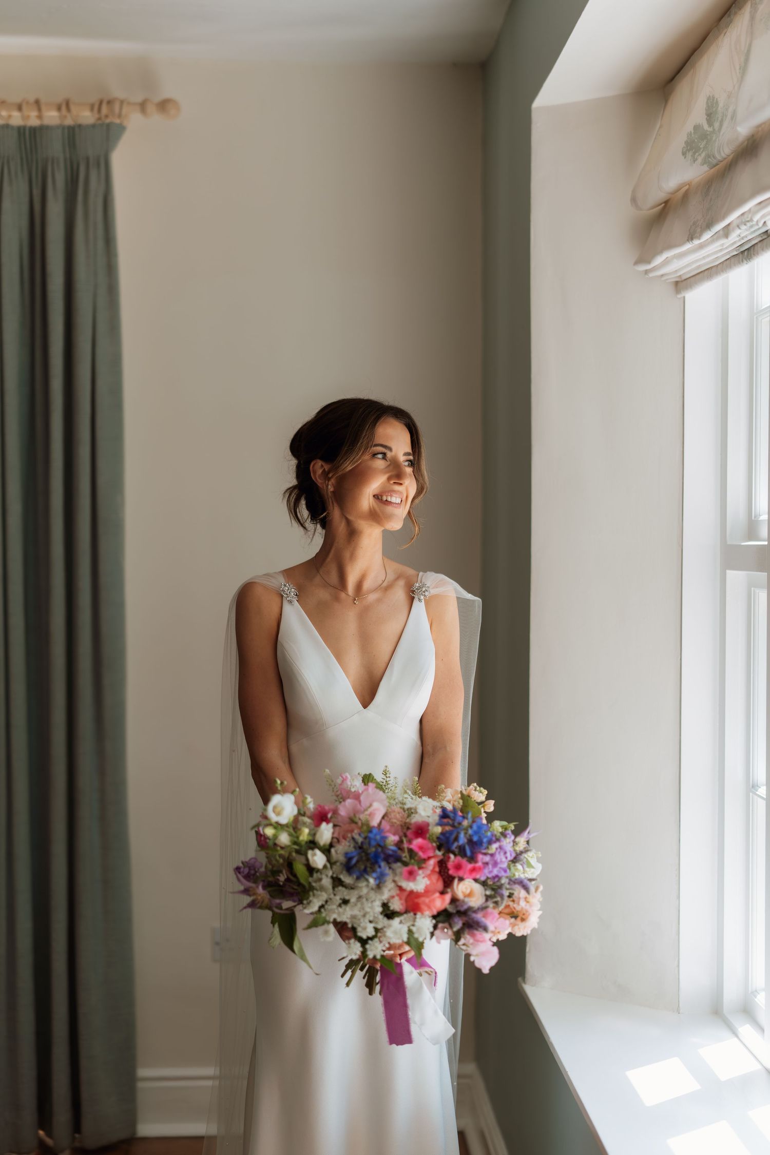 Wedding dress detail with colorful floral bouquet near window with sage green curtains.