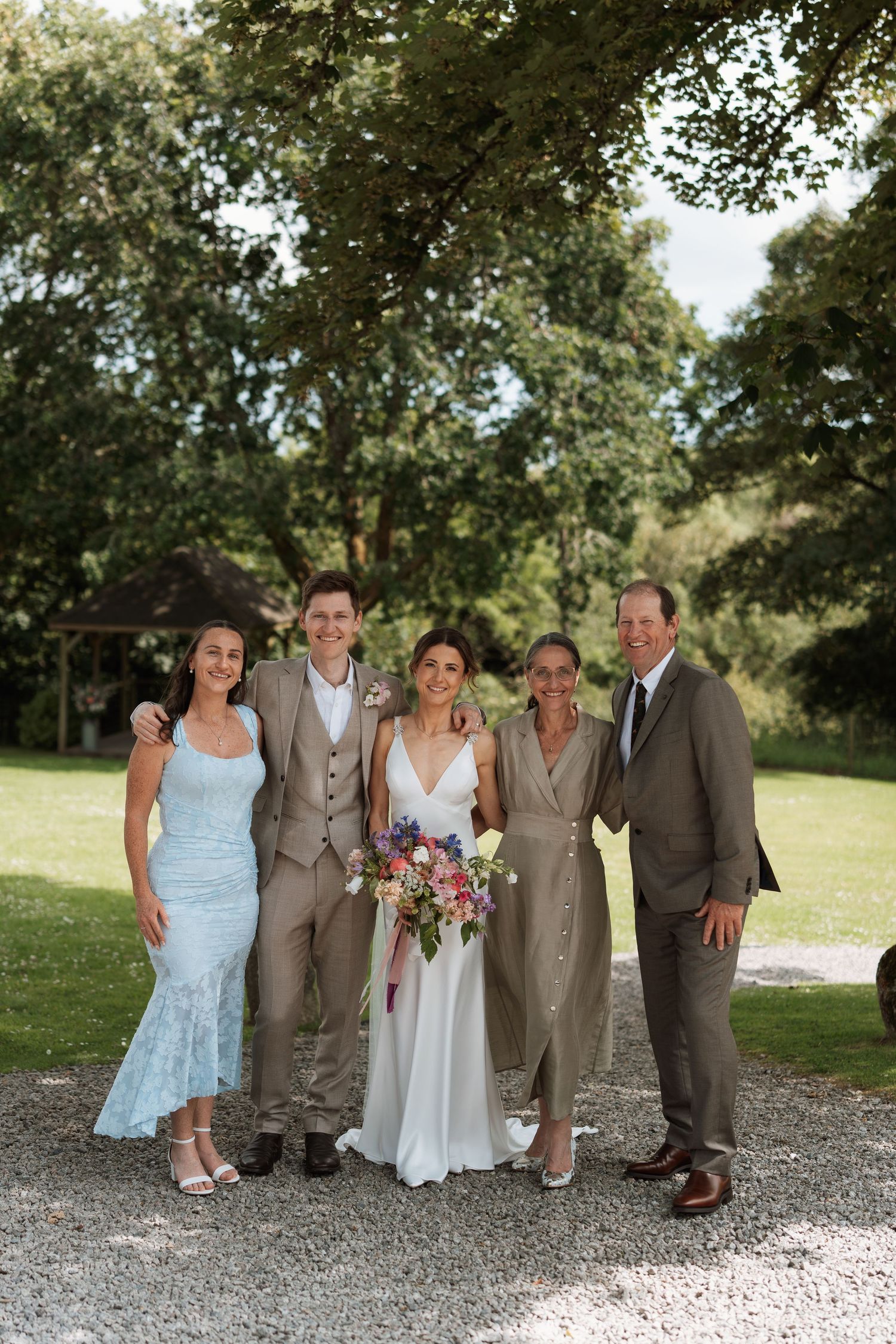 Wedding party poses together on a gravel path under green trees wearing brown suits and light dresses.