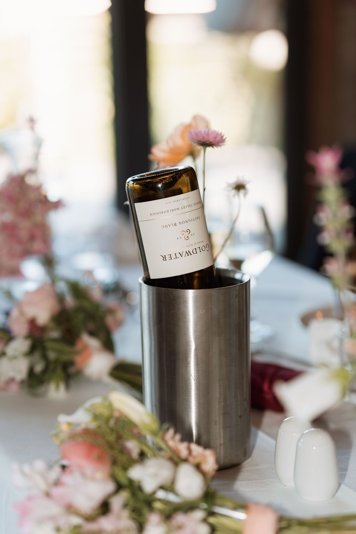 A wine bottle chills in a stainless steel cooler surrounded by delicate pink floral arrangements on an elegant table setting.
