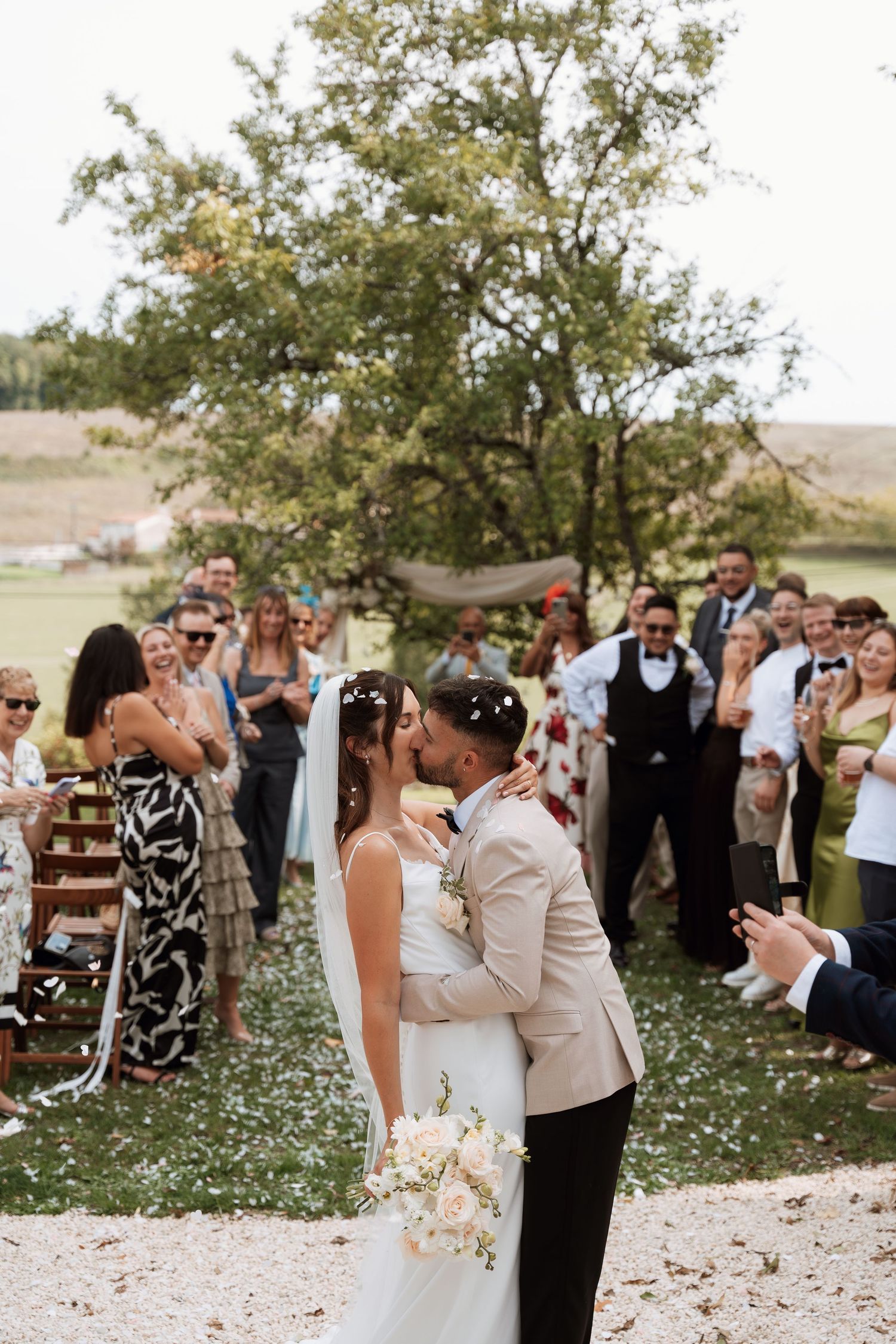 Wedding party and guests gather outdoors under a tree with white petals floating in the air.