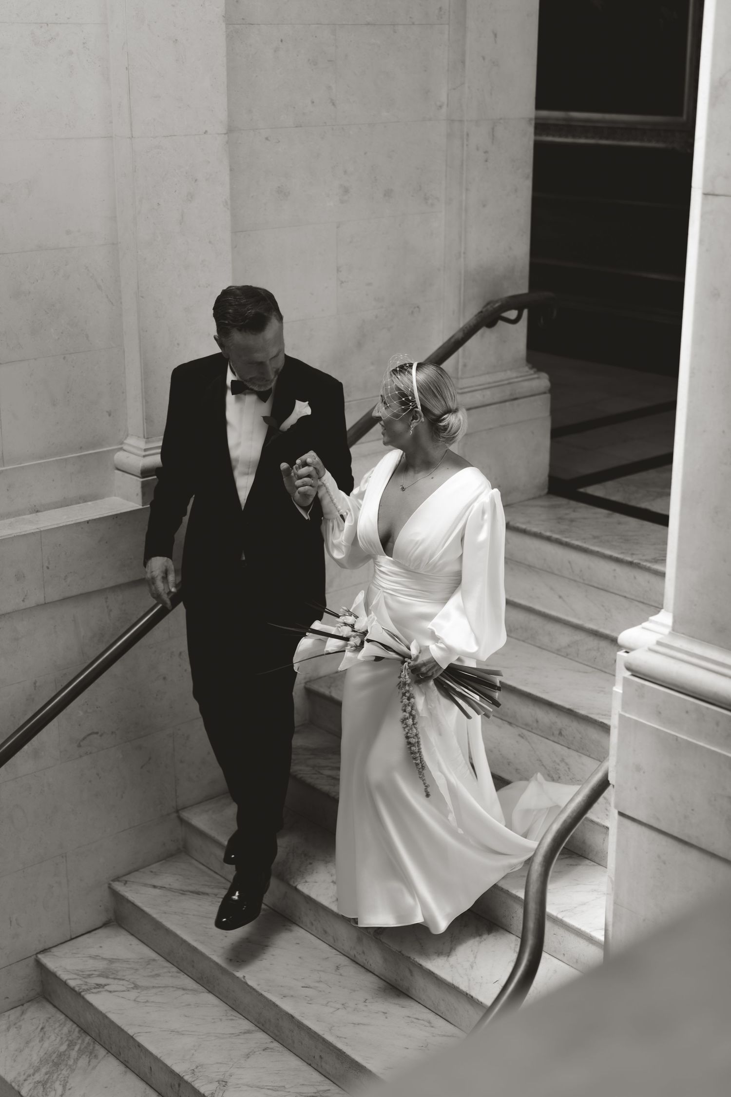 Black and white photo of a couple descending marble steps in formal wedding attire.