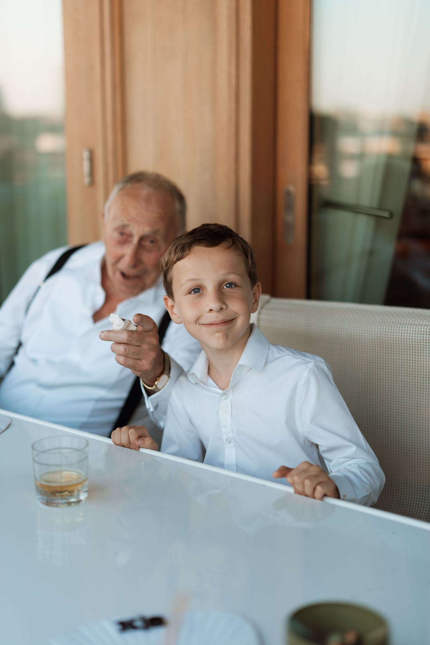 Two people sit at a white table in a dining area with a glass window behind them.