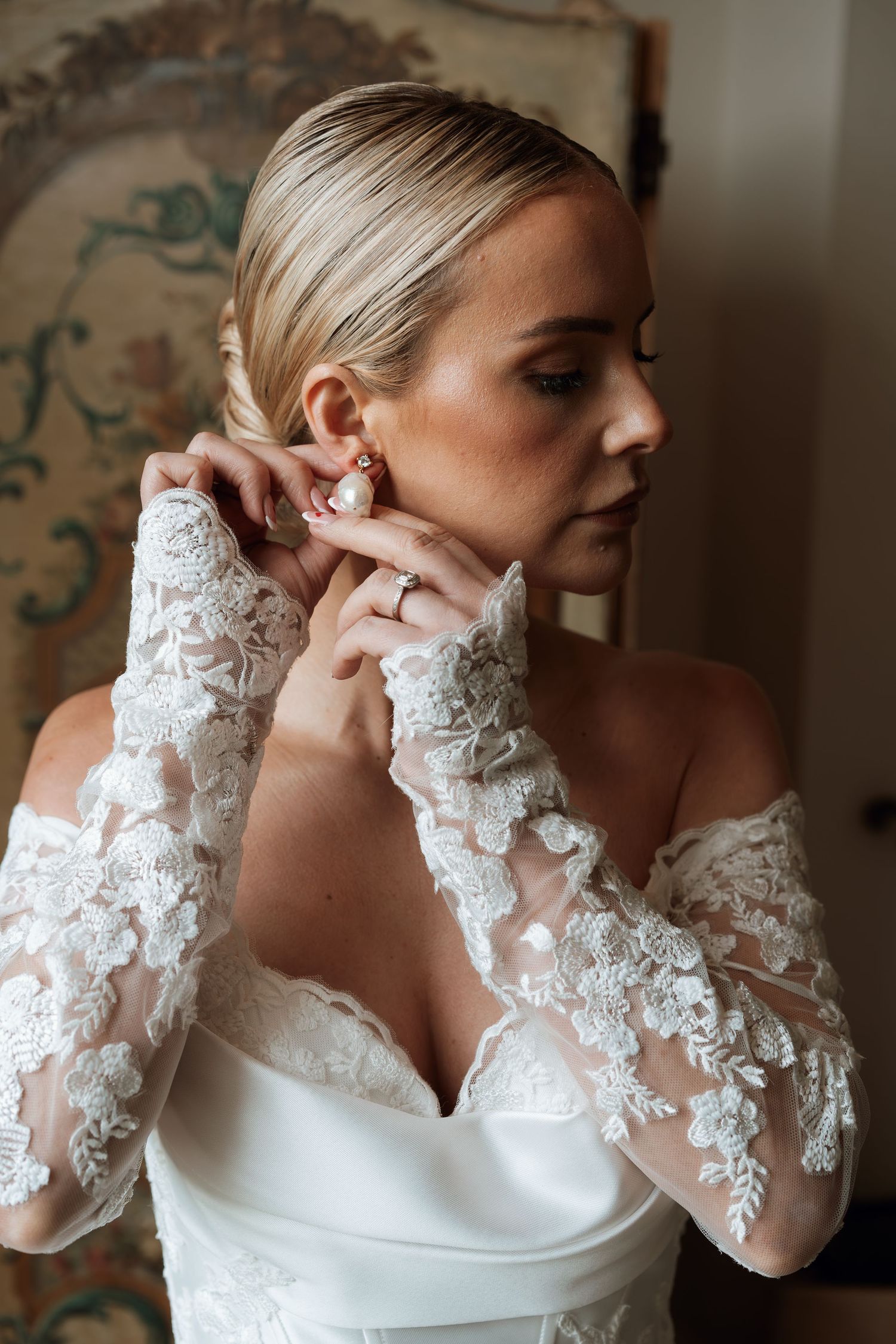Bride in lace off-shoulder wedding dress adjusting pearl earring, hair in elegant updo.