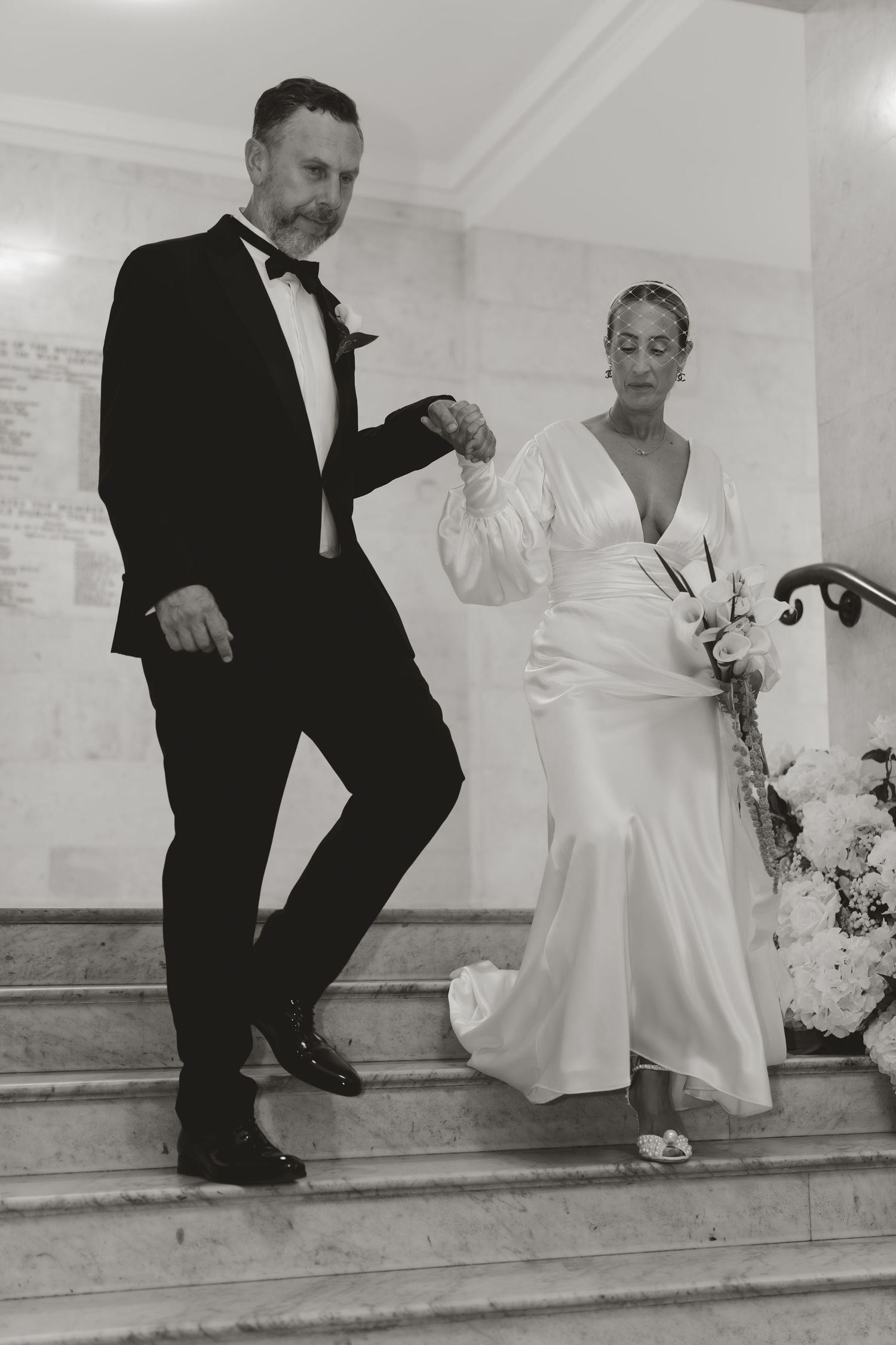 A couple descends elegant stairs at a formal event, with the bride in a flowing white dress and groom in classic tuxedo.