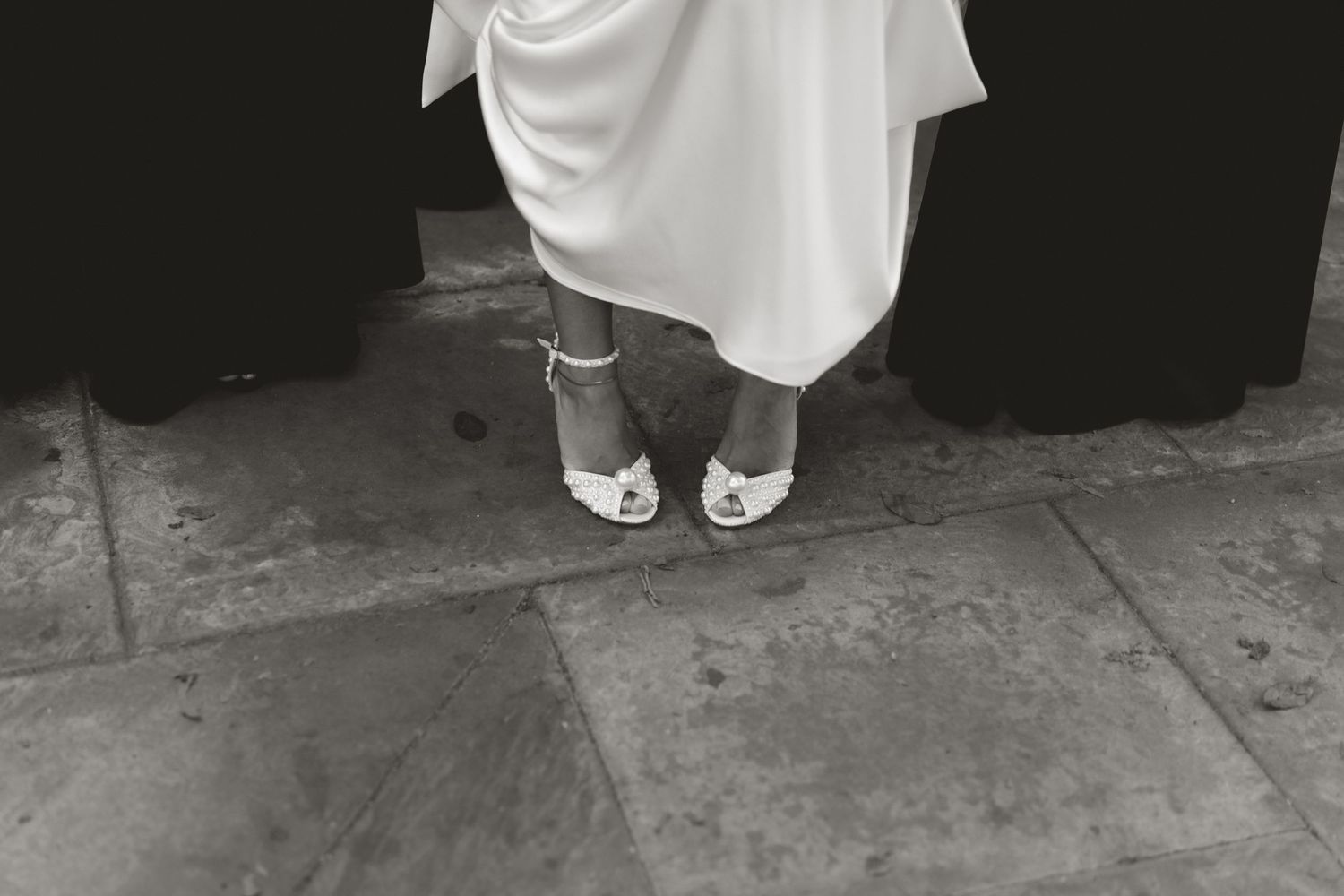 Black and white photo of white wedding shoes peeking out from under a flowing dress hem.