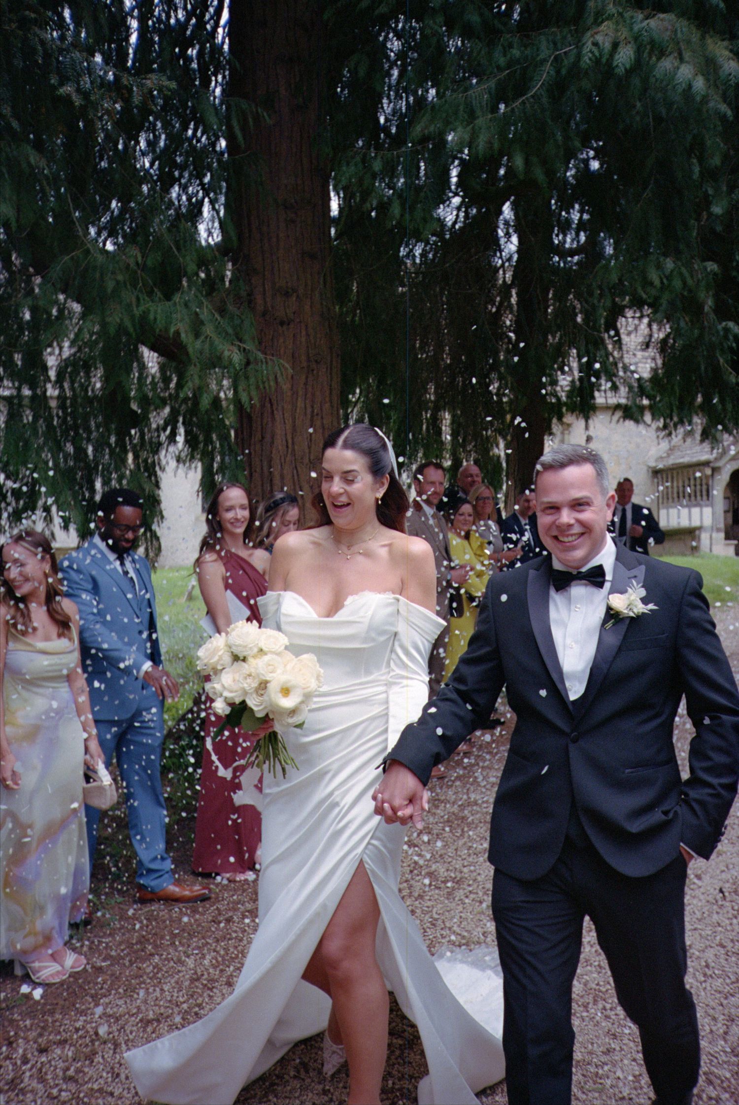 Wedding party members run and laugh together on a tree-lined path covered in fallen leaves during autumn celebrations.