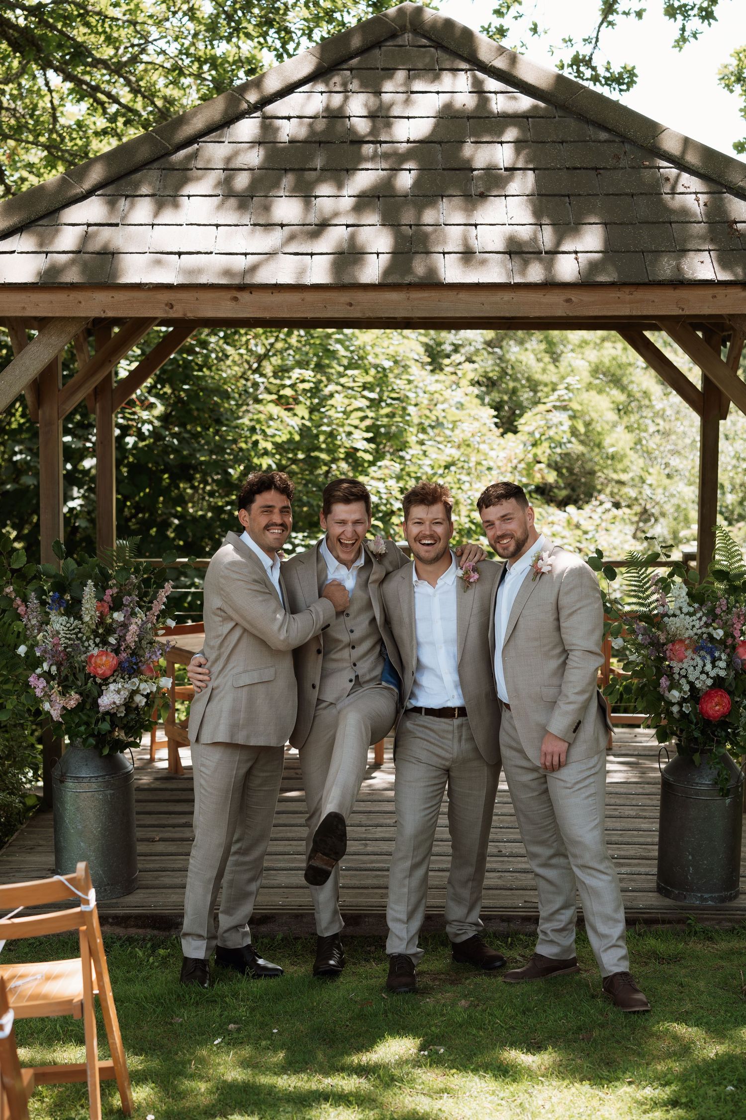 Groomsmen in beige suits pose together under a wooden gazebo decorated with floral arrangements for a summer wedding.