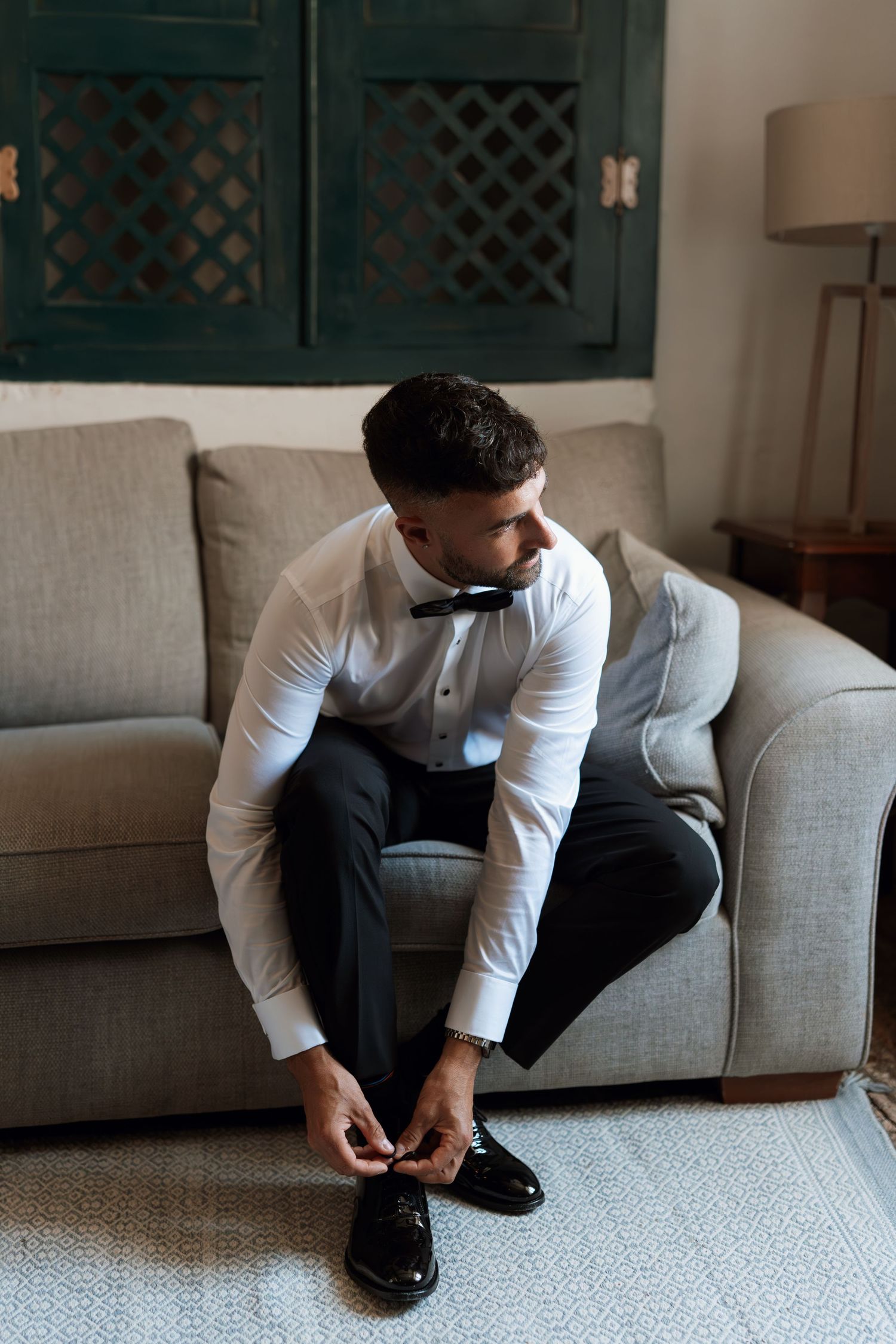 Person in formal attire sitting on a gray couch tying dress shoes.