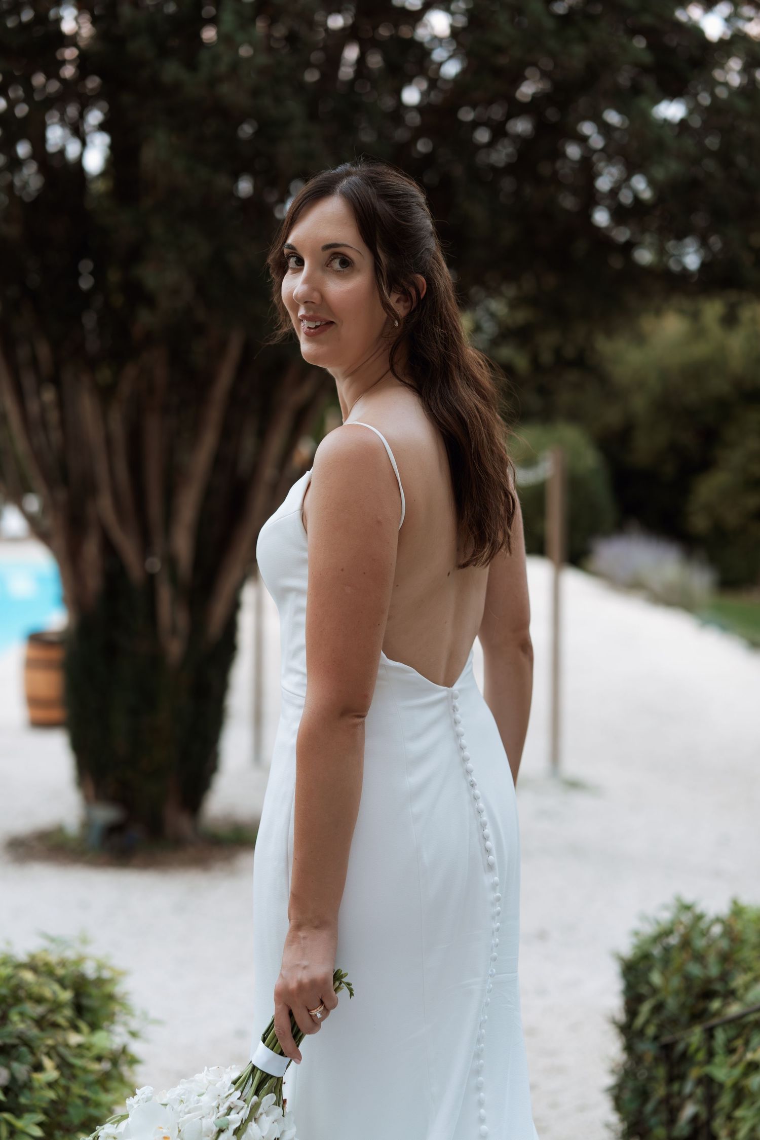Bride in elegant white dress poses outdoors among trees and greenery.