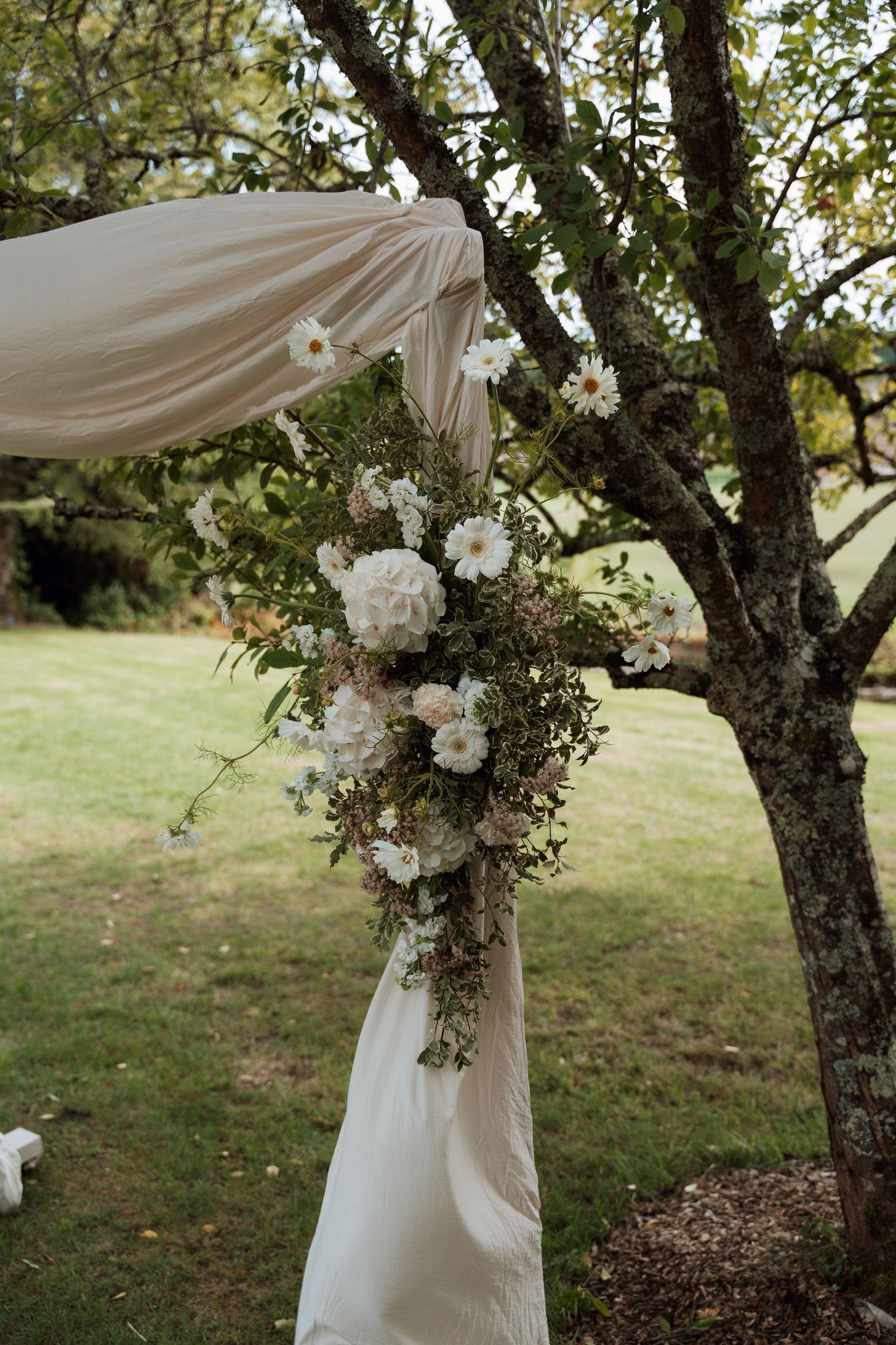White floral arrangements and draped fabric decorate tree branches in an outdoor wedding ceremony setting.