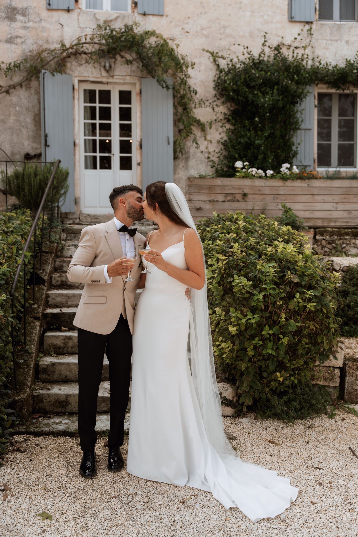 Couple in wedding attire sharing a private moment on gravel path by stone building with blue shutters.