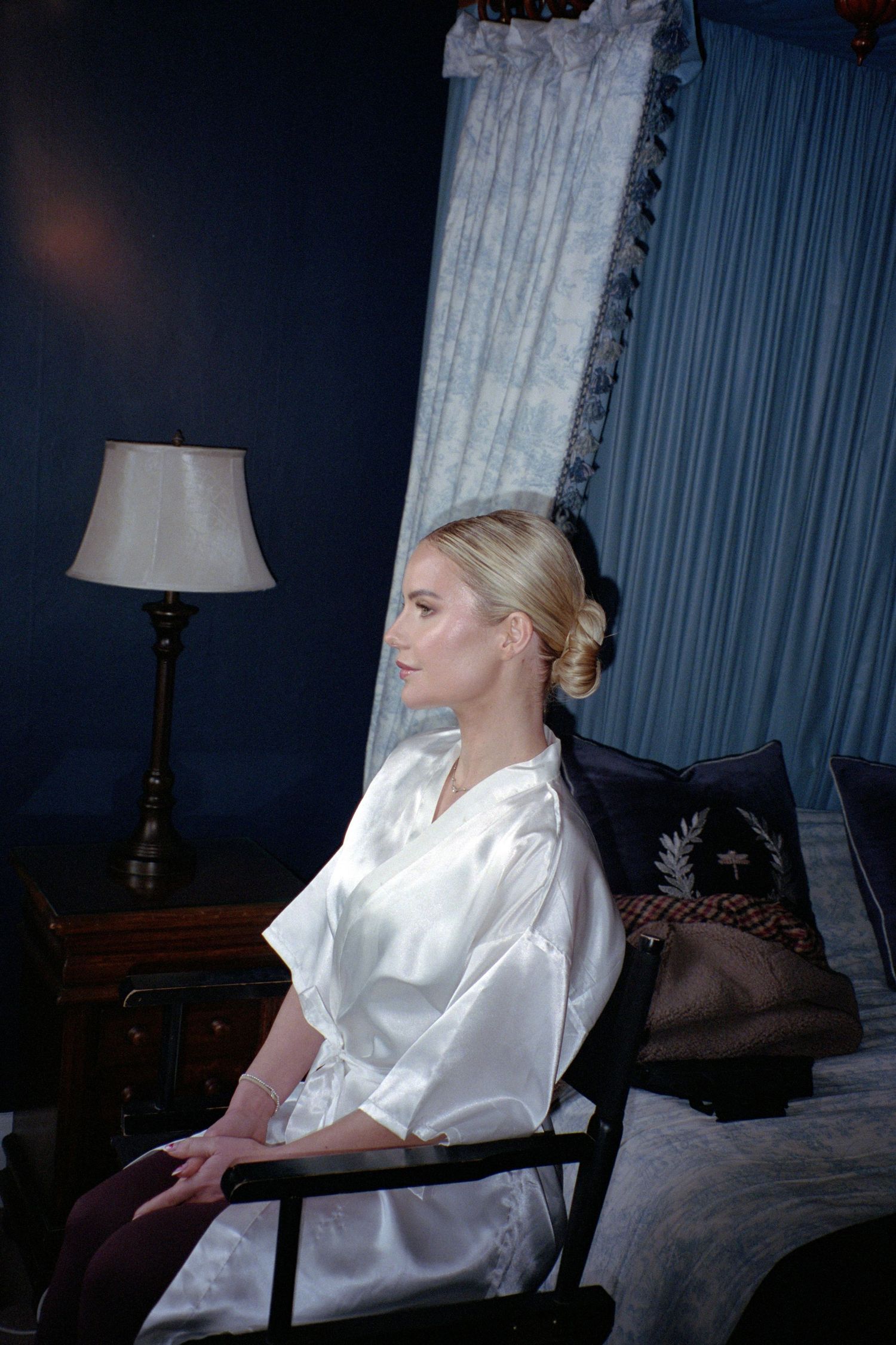 A figure in a white silk robe sits in a dark room with curtains and a table lamp creating a moody atmosphere.