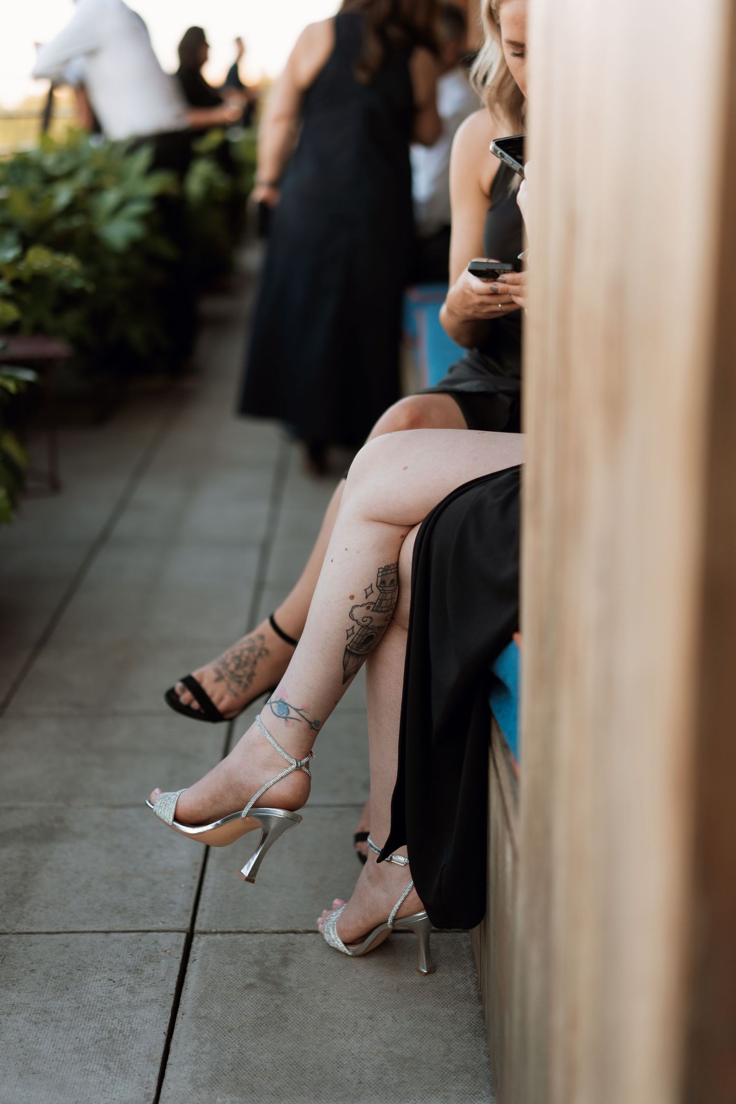 Close-up of tattooed legs in silver heels resting on outdoor seating.