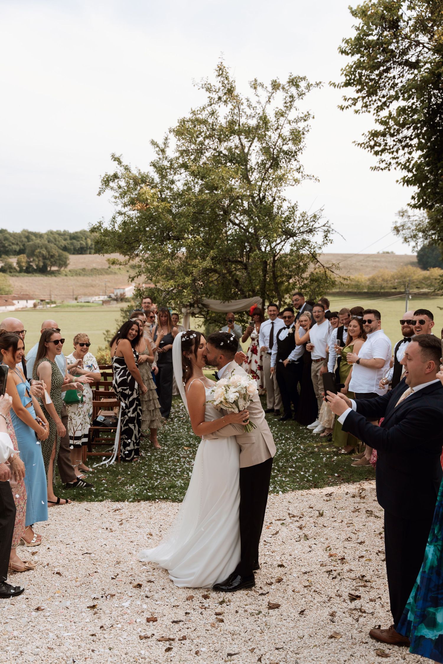 Wedding guests gather outdoors for a ceremony on a sunny day with trees and greenery in the background.