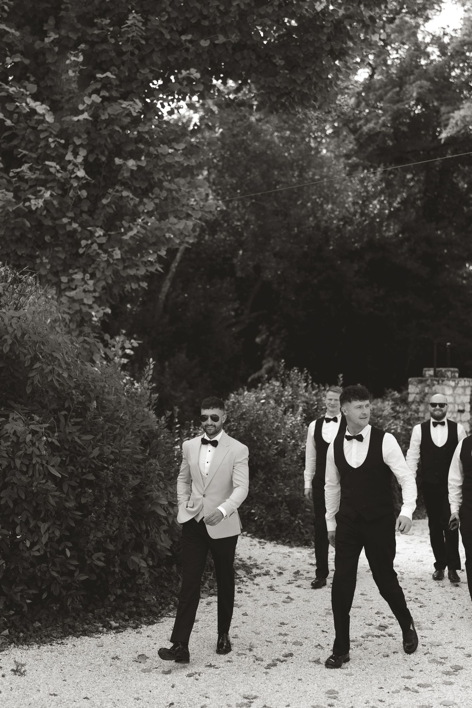 Group of formally dressed people walking in a line through a garden setting in black and white.