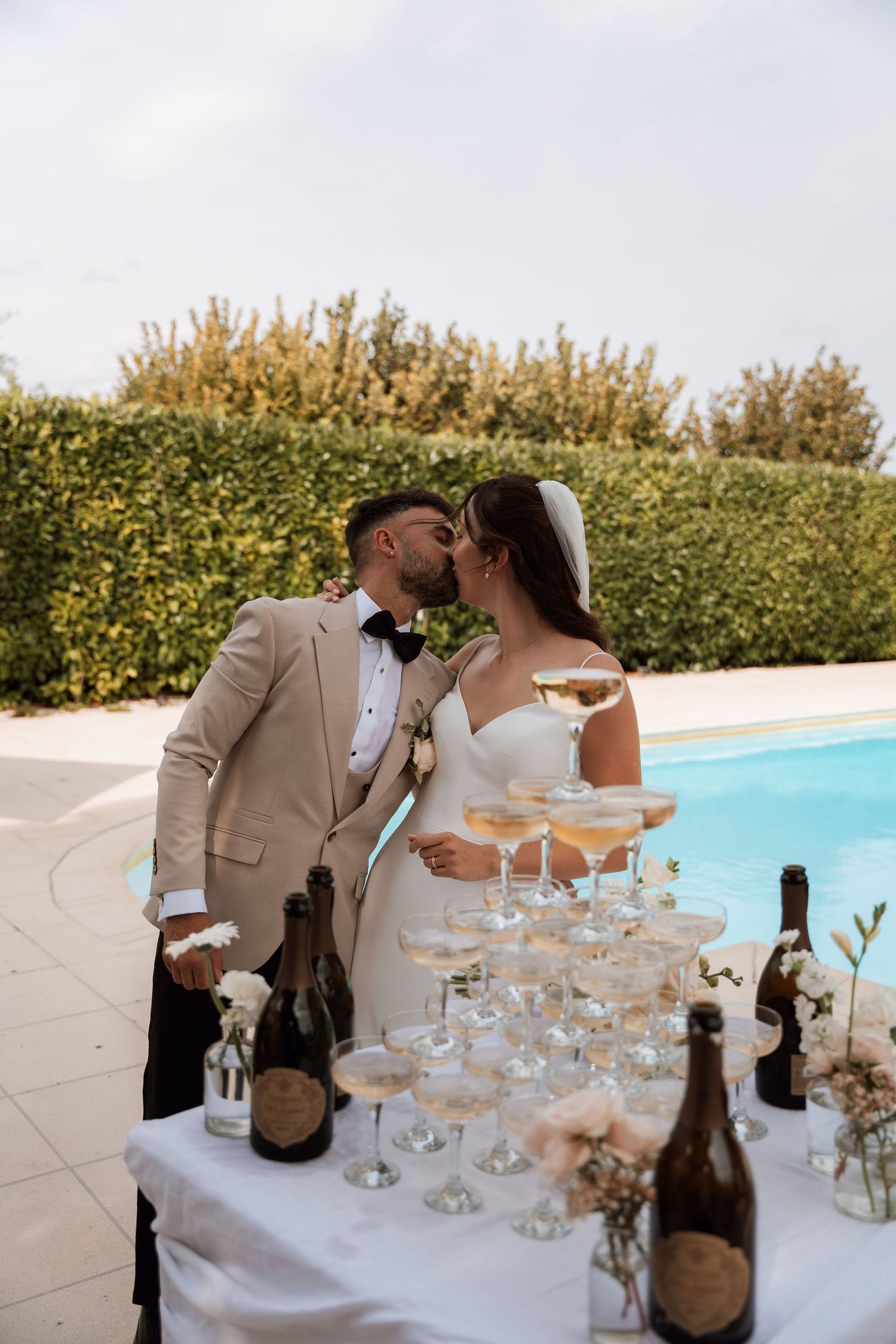 A champagne tower sits on a white tablecloth beside a swimming pool at an elegant wedding reception.