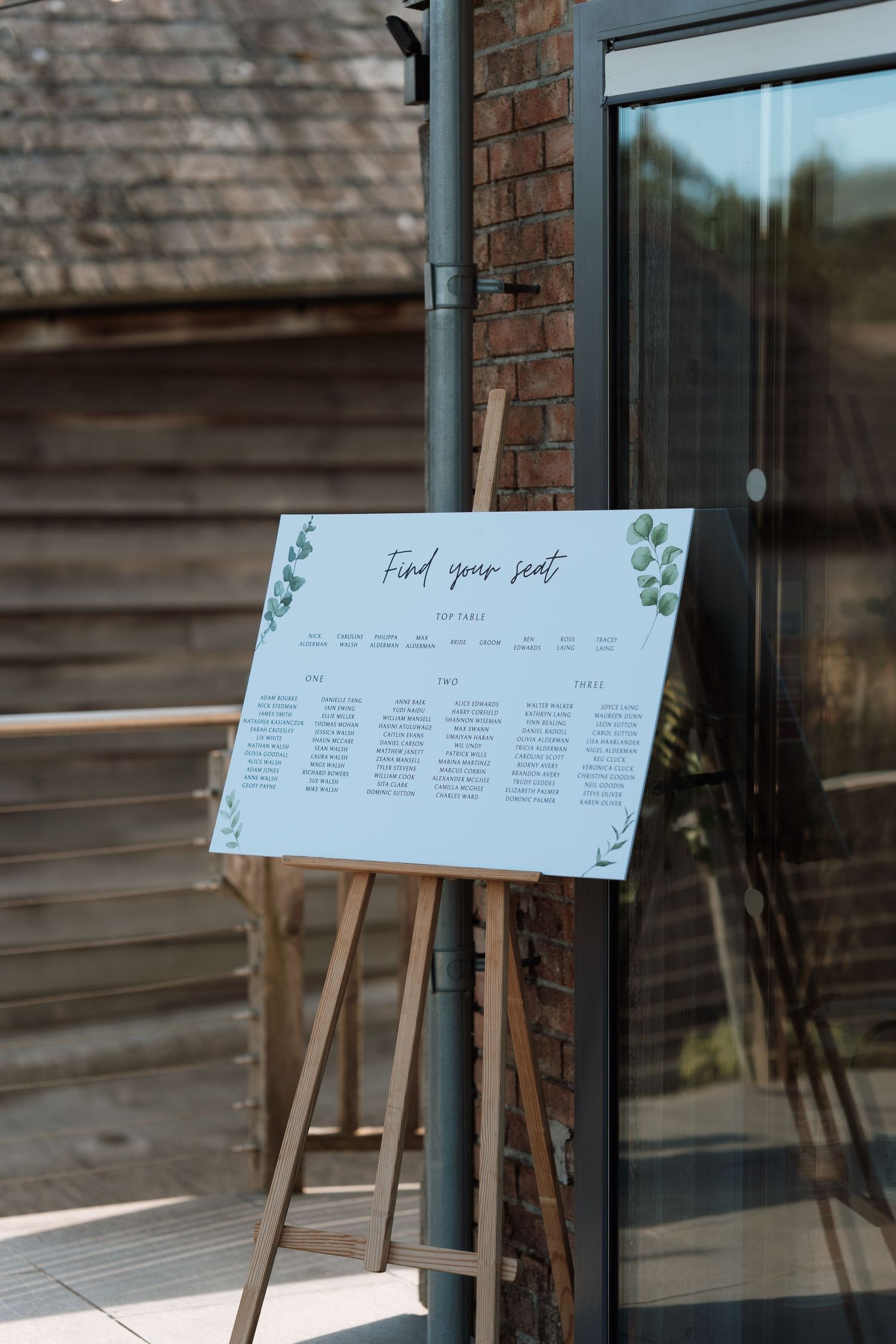 Wedding seating chart displayed on a wooden easel against a brick wall with greenery accents.