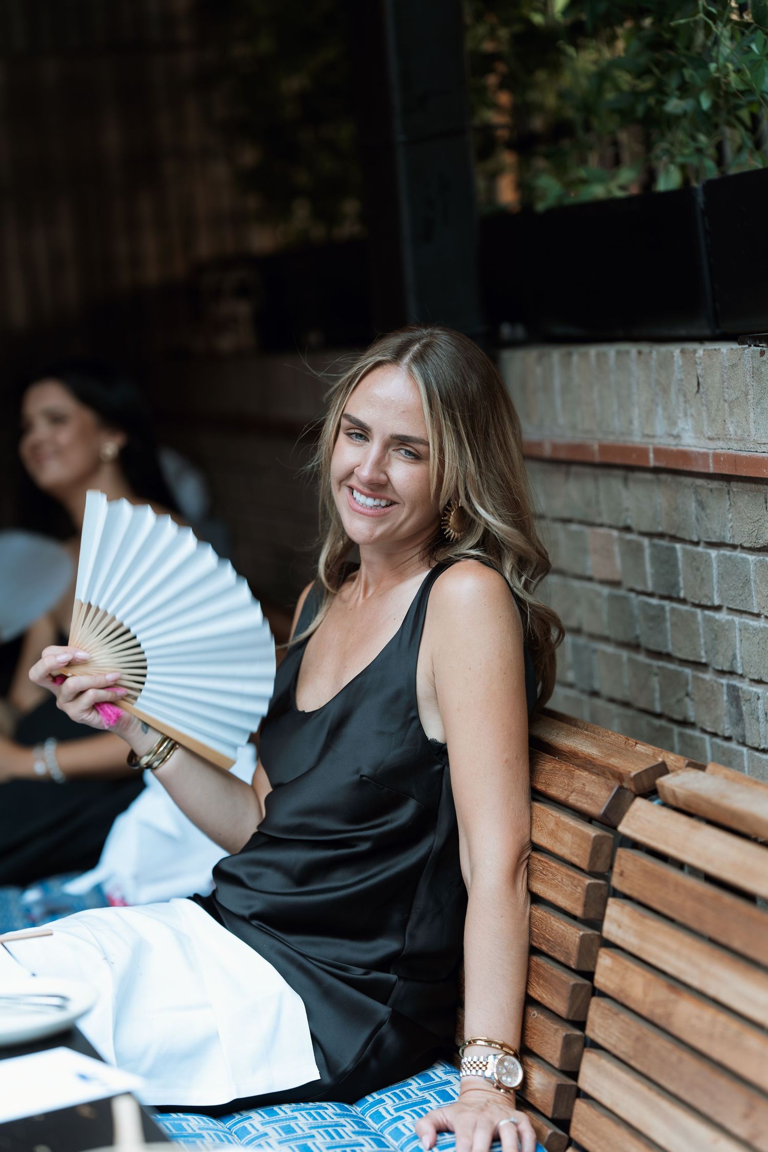 A person in a black dress sits on a wooden bench holding a decorative white folding fan.
