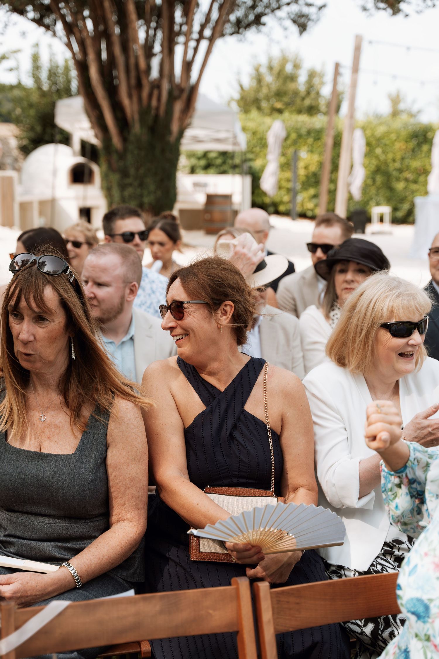 Wedding guests seated outdoors in white chairs smile and laugh during a ceremony.