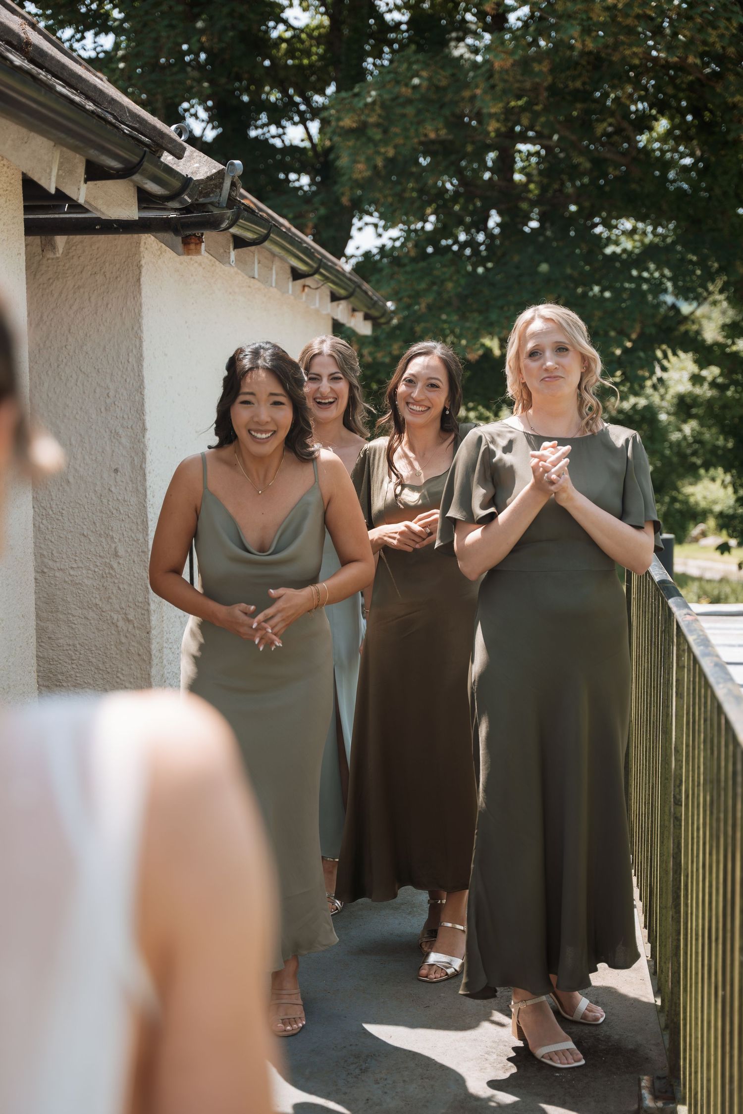 Bridesmaids in olive green dresses share joyful moments on a sunny outdoor patio.