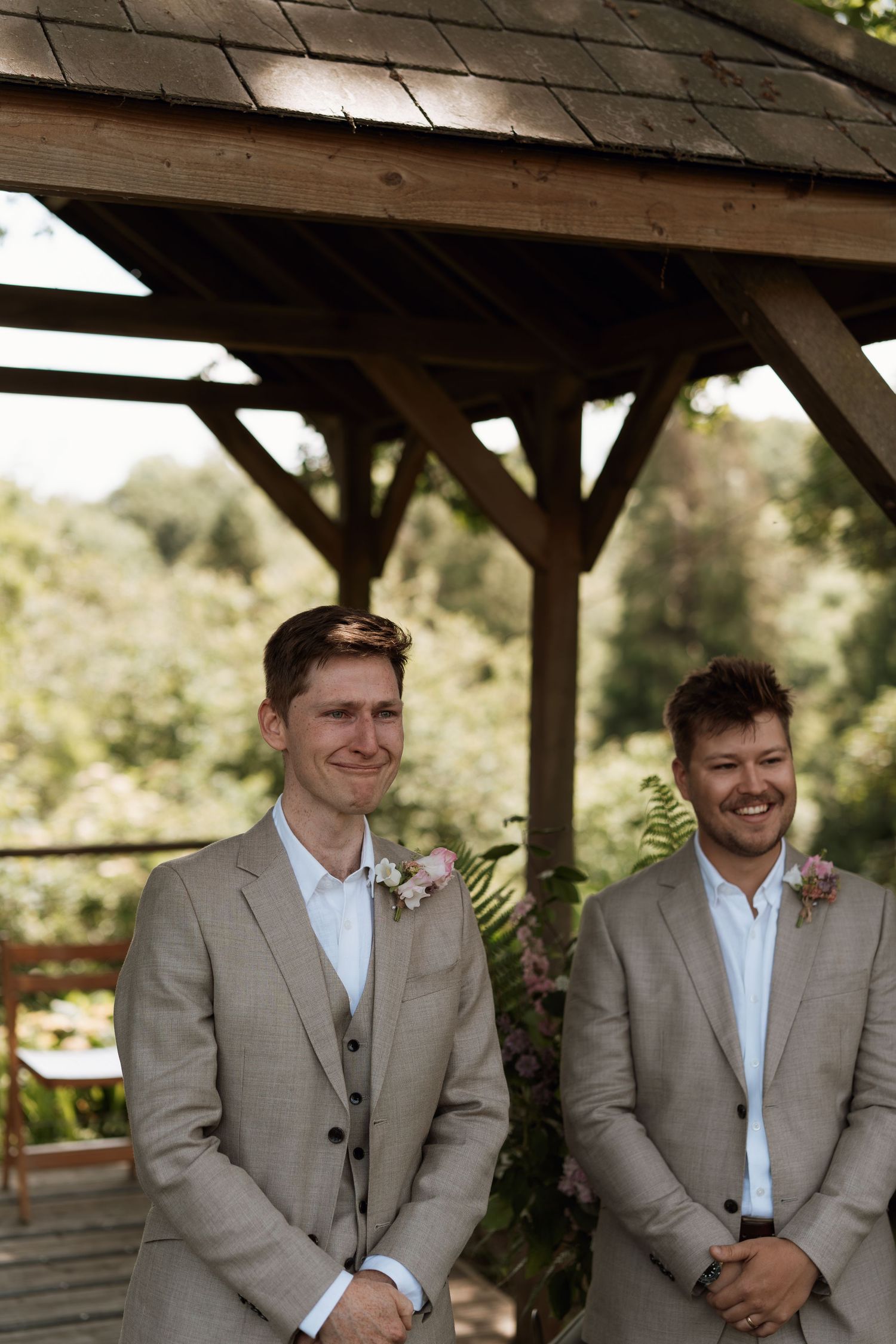 Groomsmen in light beige suits standing together under a wooden pavilion.