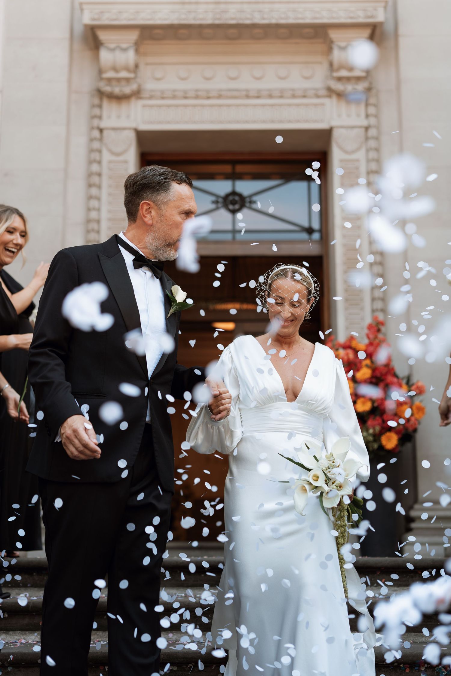 Wedding party exits venue as white confetti rains down around them.