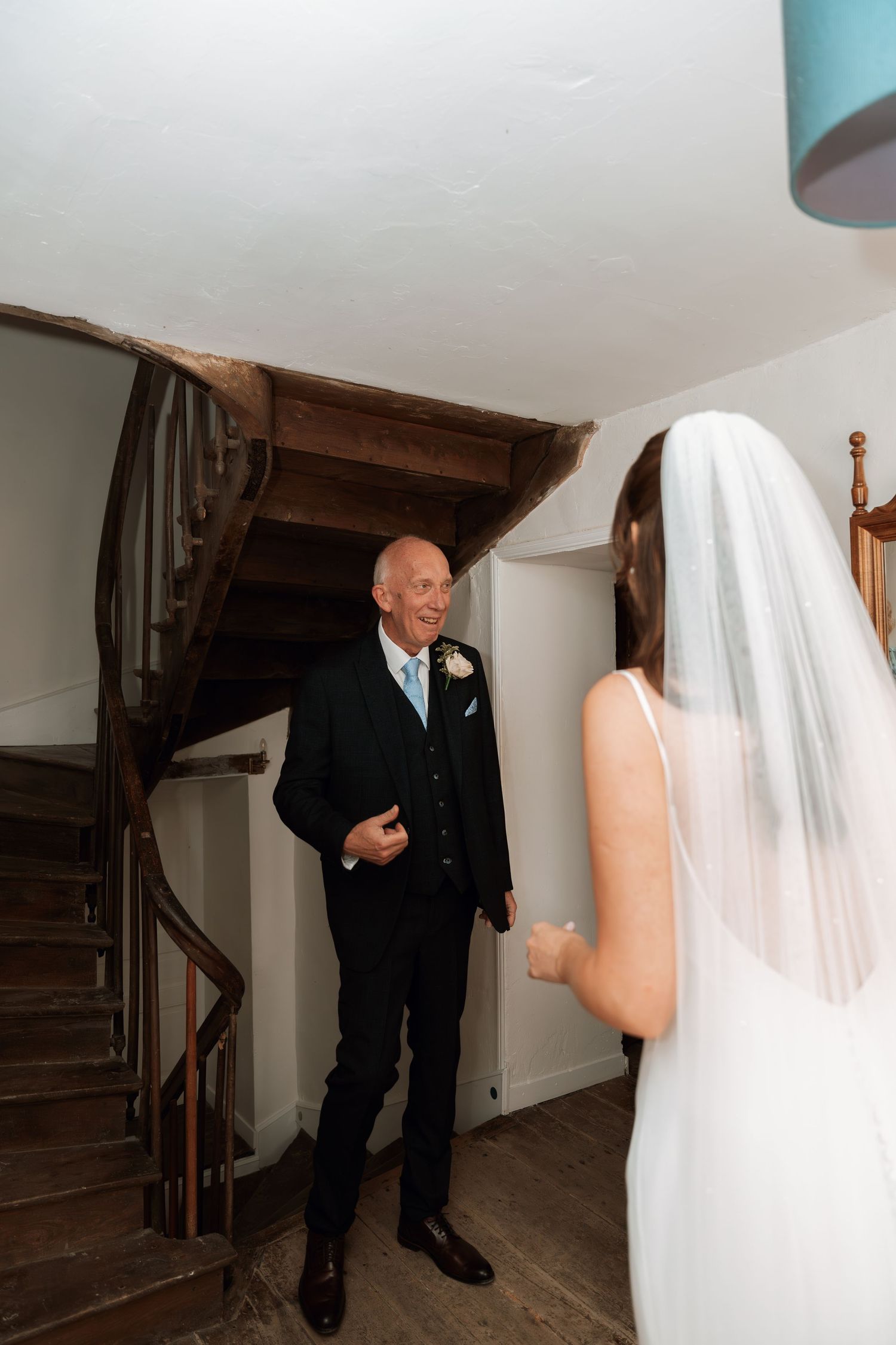 First look moment between bride in white gown and suited figure near wooden staircase.