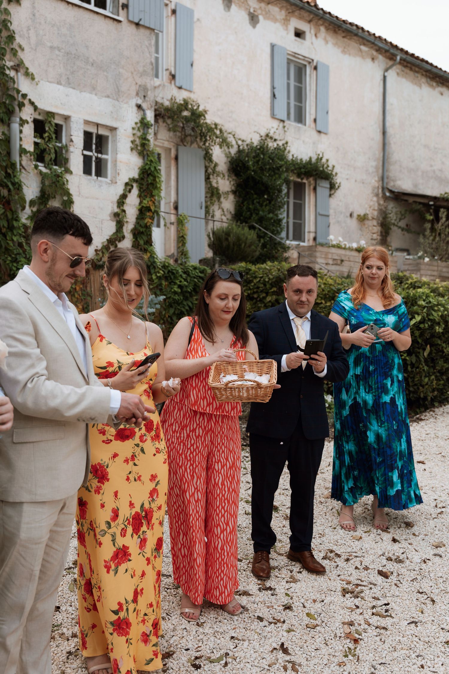 Wedding guests in colorful floral dresses gather outside a rustic French chateau.