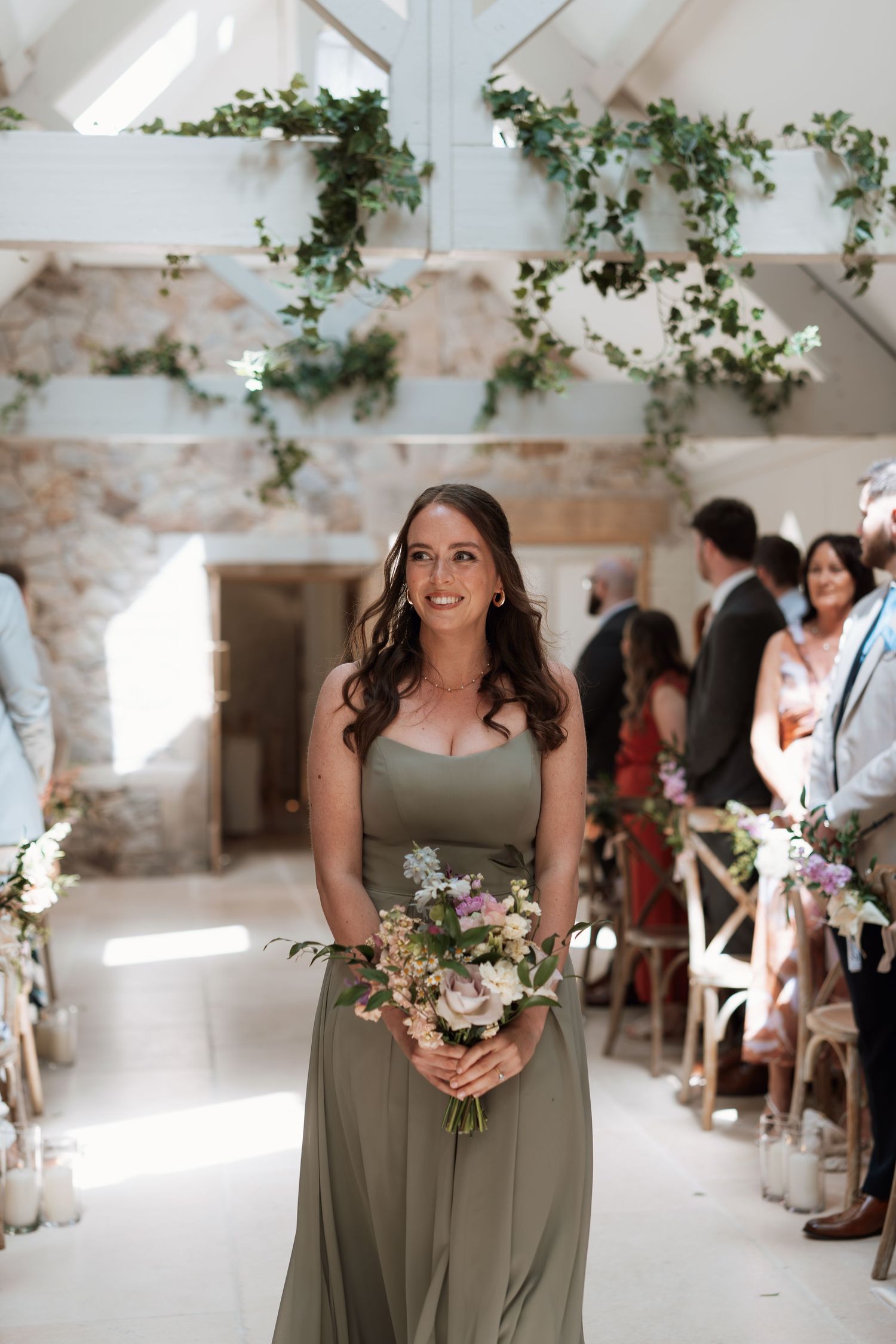 Bridesmaid in sage green dress walks down aisle holding floral bouquet in rustic stone wedding venue.