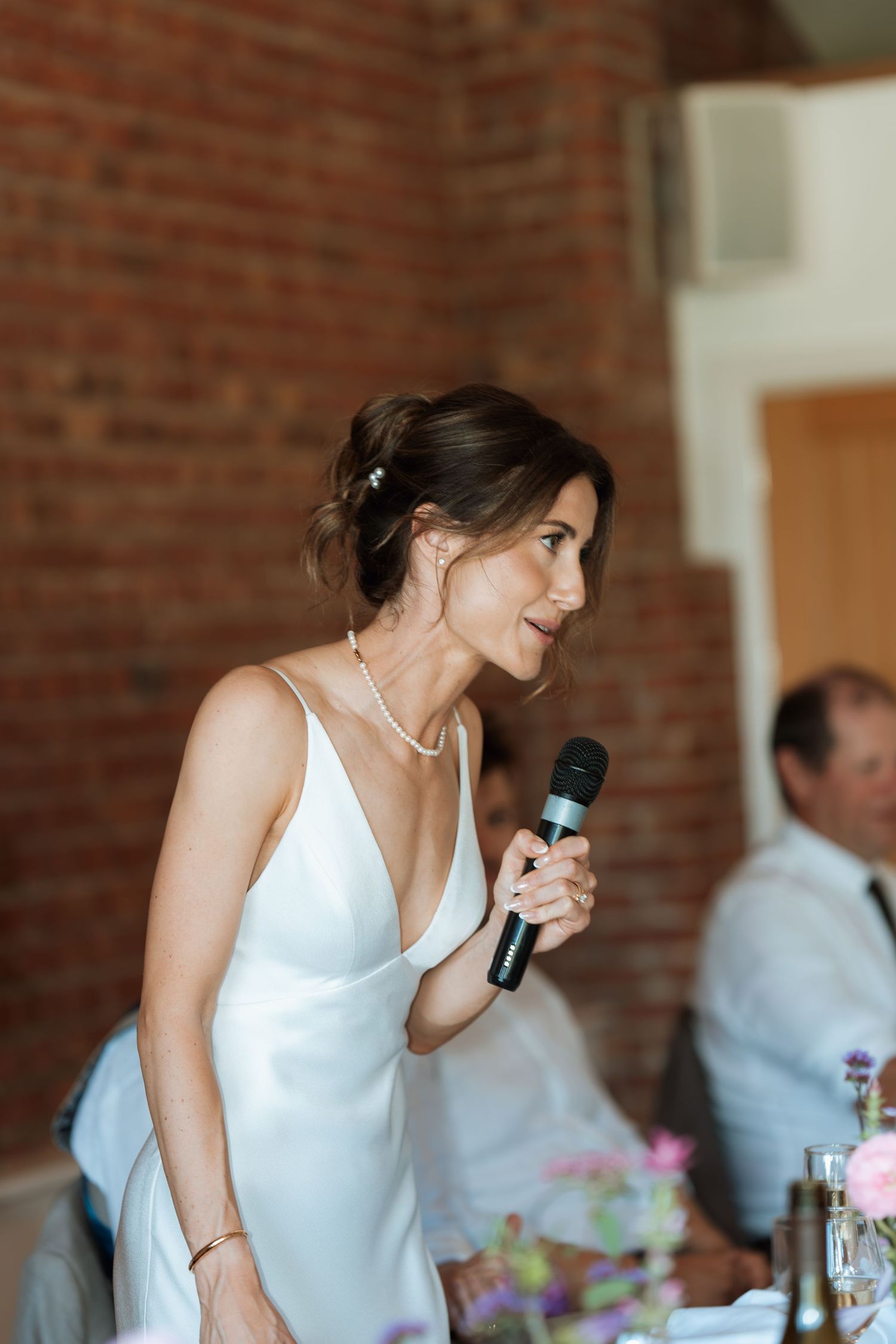 Bride giving speech with microphone at reception venue with exposed brick walls.