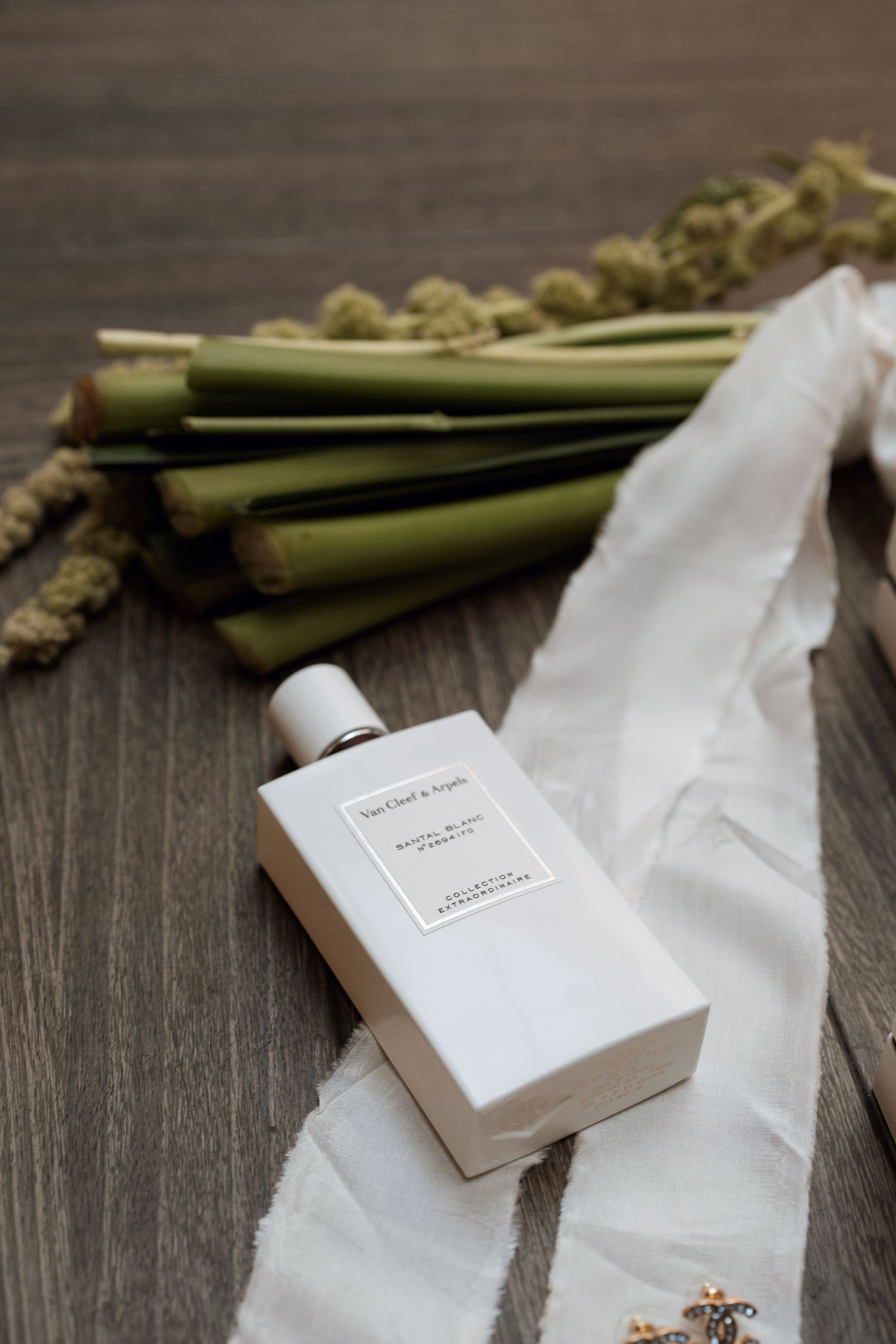 White perfume bottle and green stems arranged on wooden surface with delicate fabric.