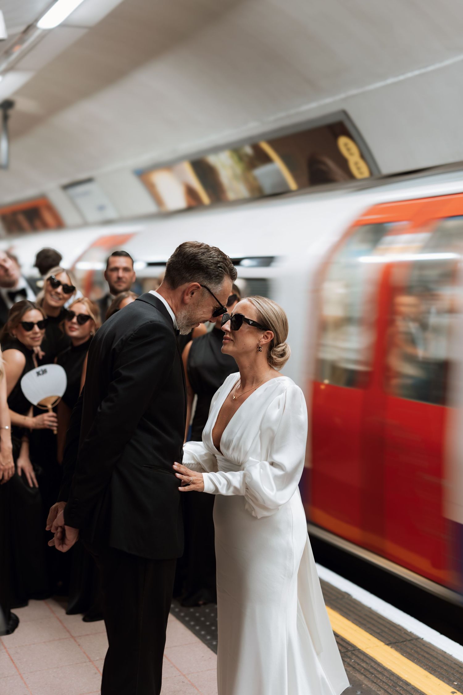 Wedding party celebrates on a subway platform with a train passing by in the background.