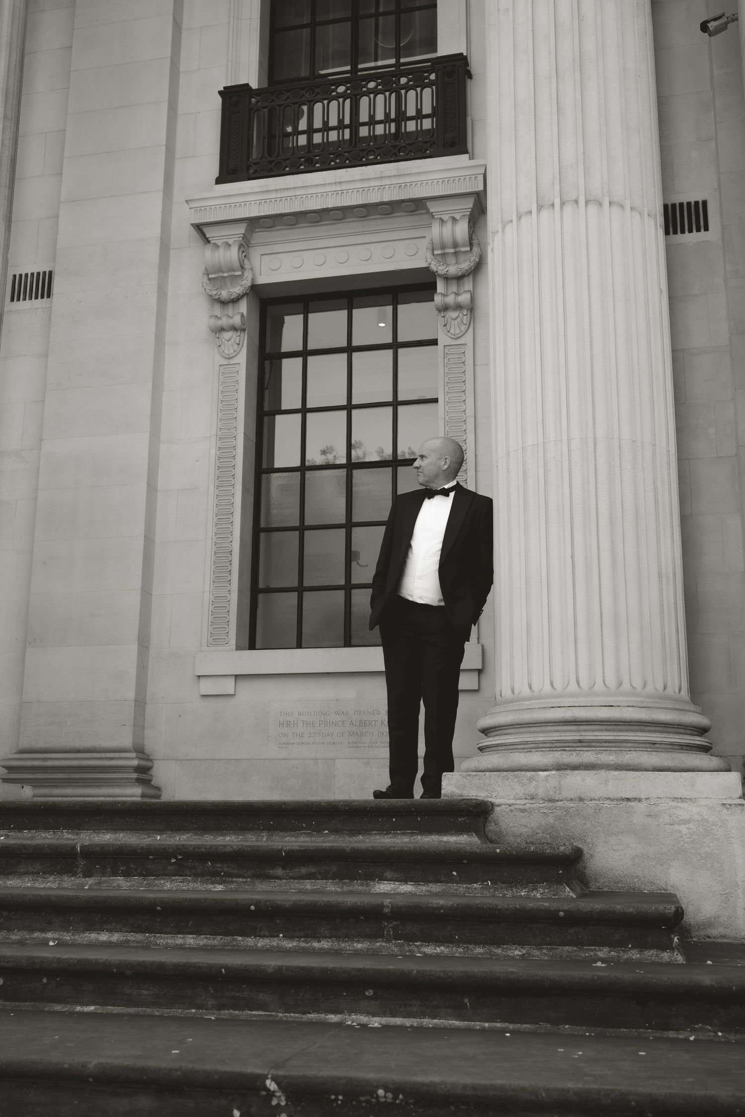 A black and white photo of a person in formal attire standing by a classical column and window of a grand building.