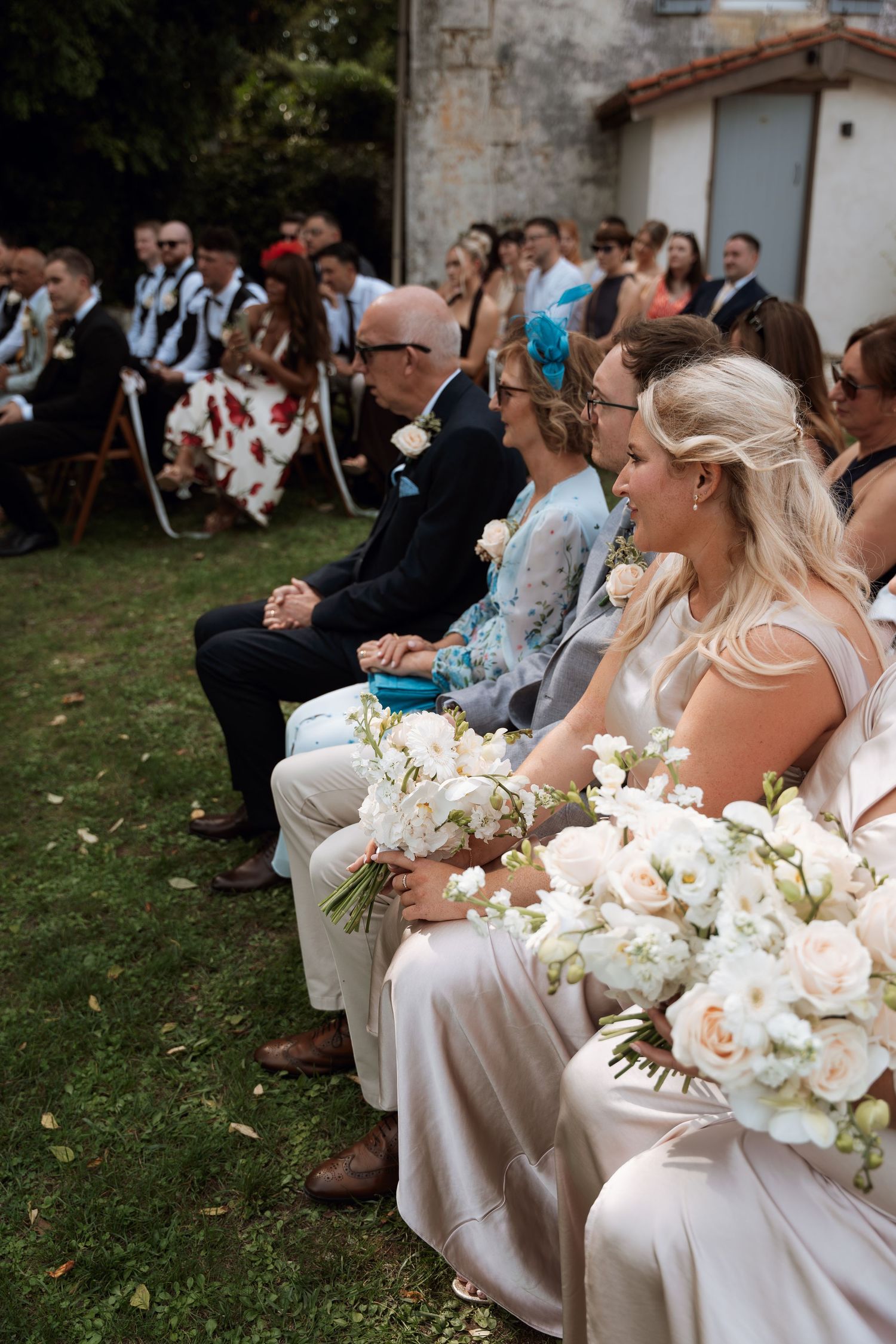 Wedding party members sit in white dresses holding elegant white floral bouquets during an outdoor ceremony.