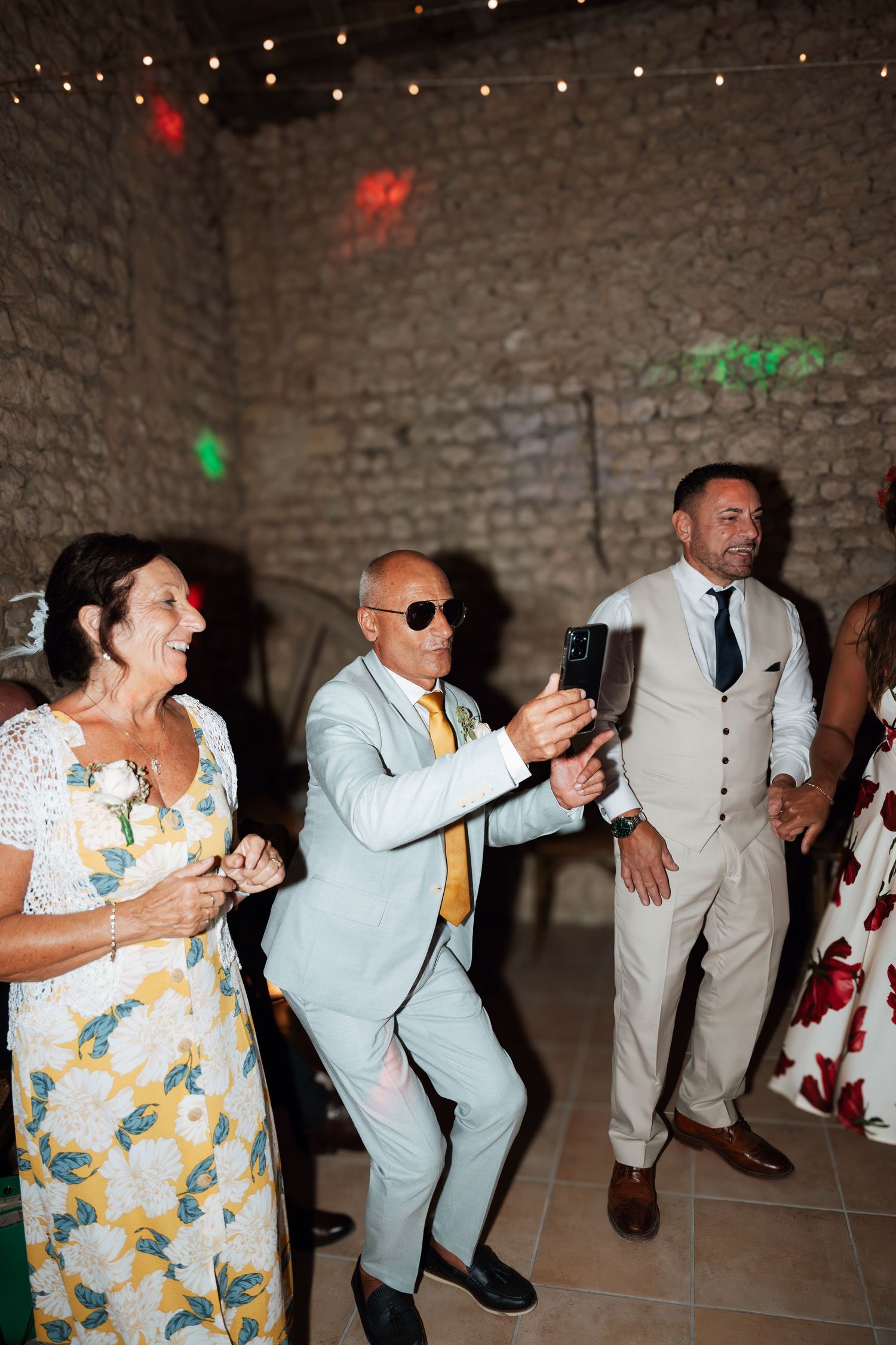 Wedding party dancing in line formation inside rustic stone venue with string lights.