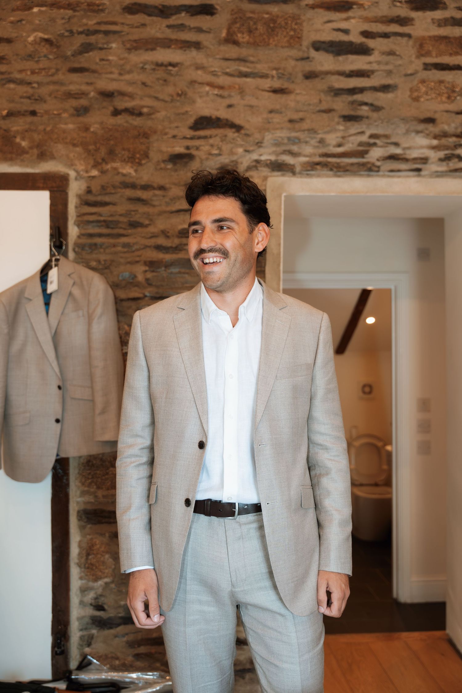 Multiple angles of a light gray suit jacket and pants hanging on display in a rustic stone-walled room.