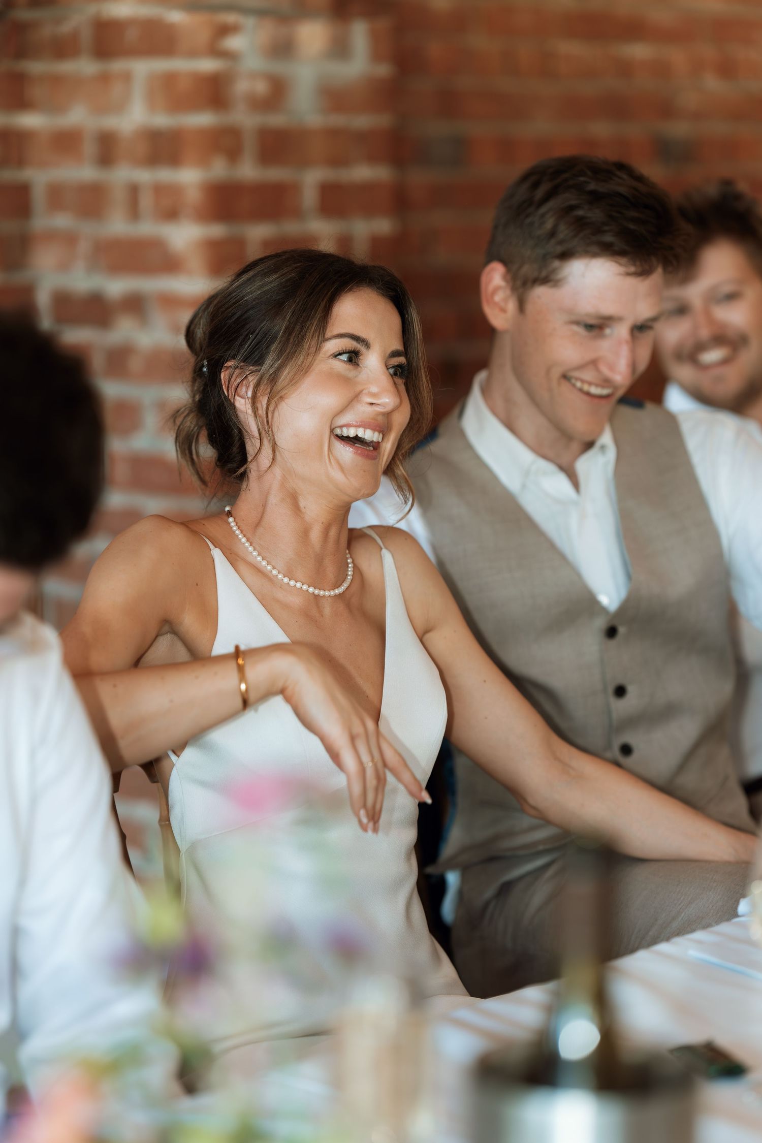 Candid reception moment showing laughter and joy at wedding table with brick wall backdrop.