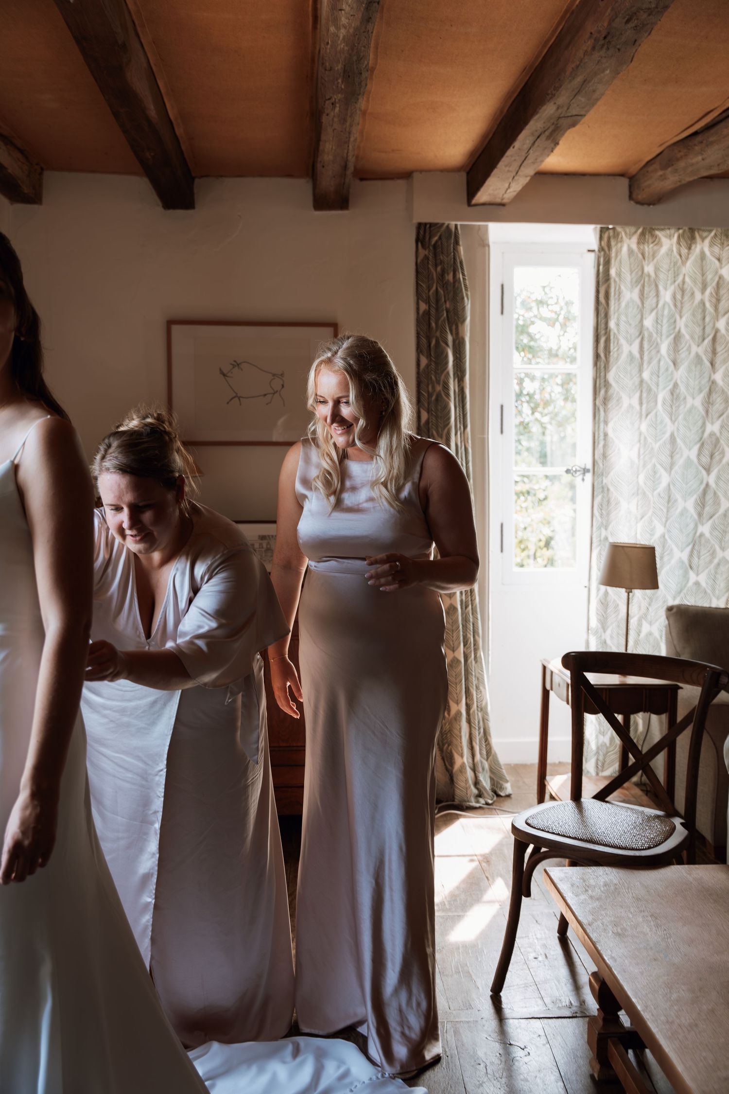 Bridesmaids in white dresses gather in a rustic room with wooden beams.