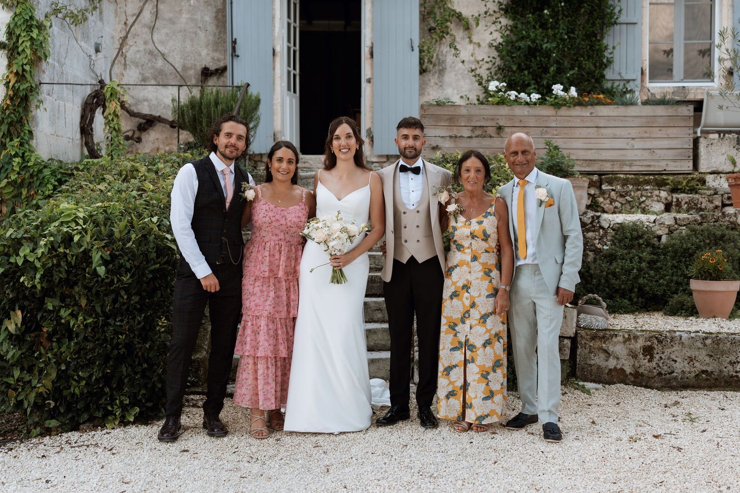 Wedding party and family members pose for formal group photos outside rustic stone building.