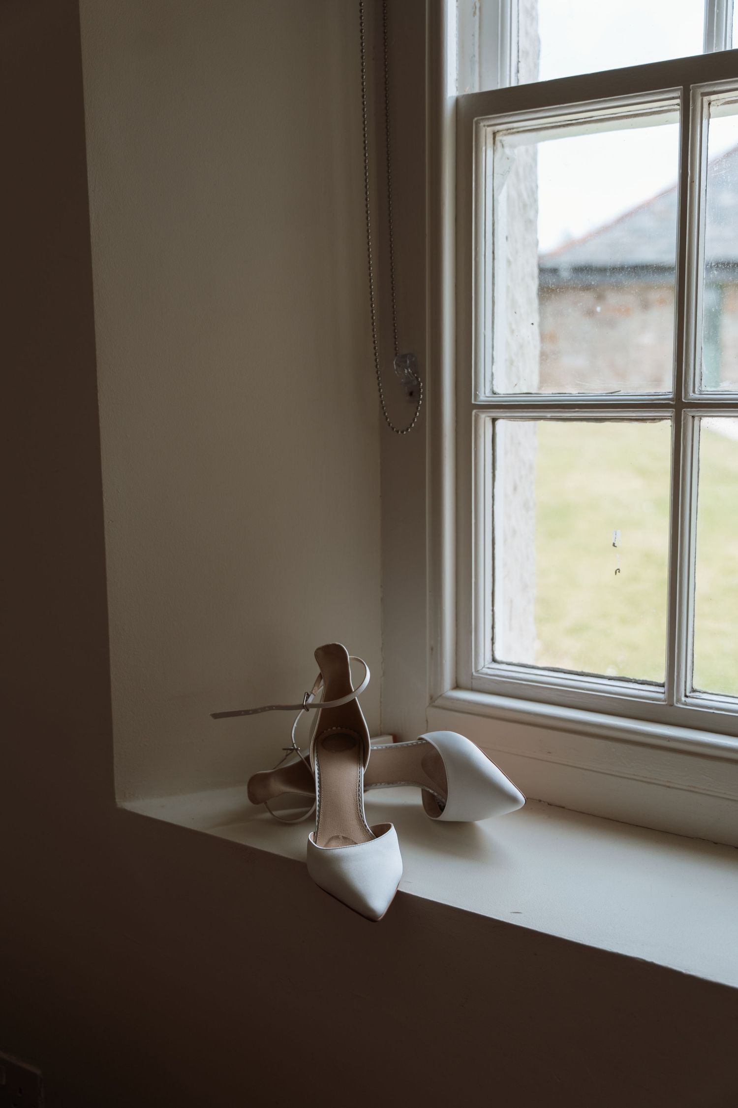 A pair of elegant white bridal shoes sits on a windowsill in natural light, creating a minimalist wedding detail shot.