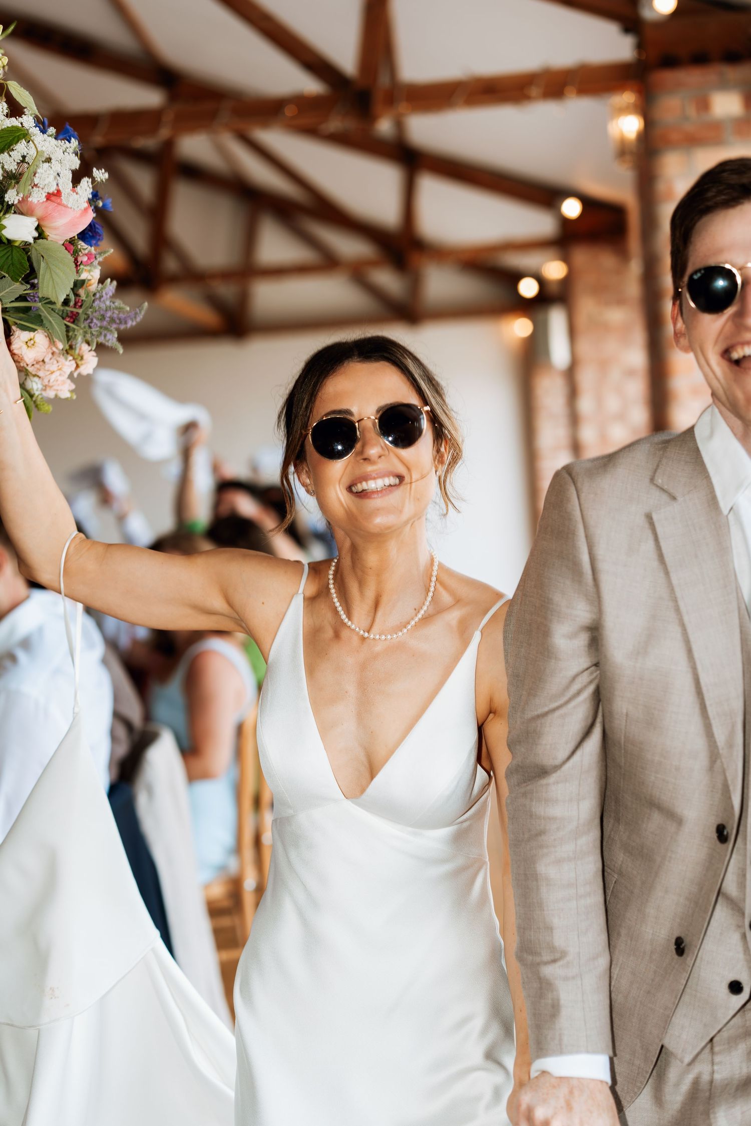 Wedding celebration with couple wearing sunglasses and holding bouquet under rustic wooden beams.