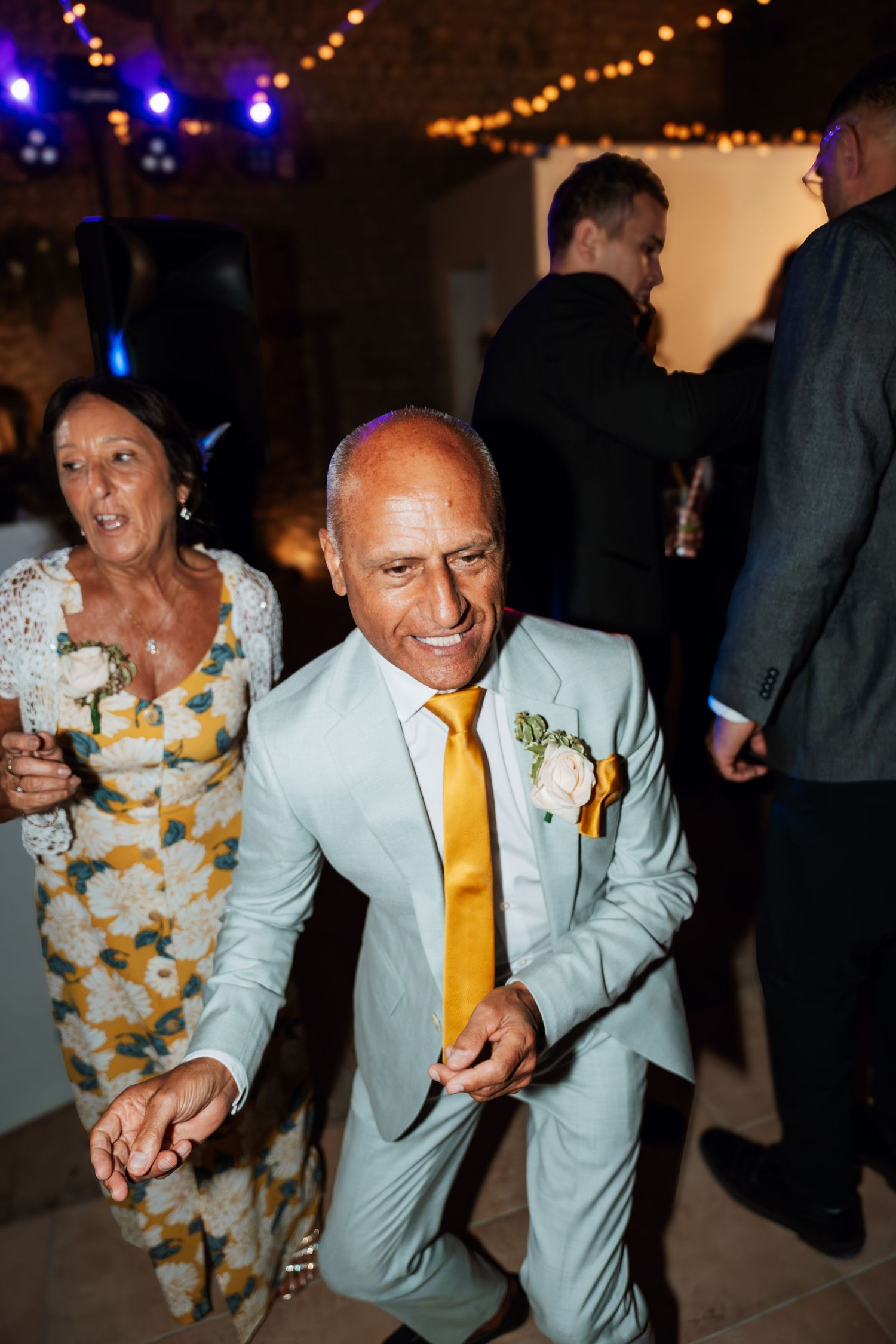 Wedding guests in light suits dancing energetically at a reception with string lights overhead.