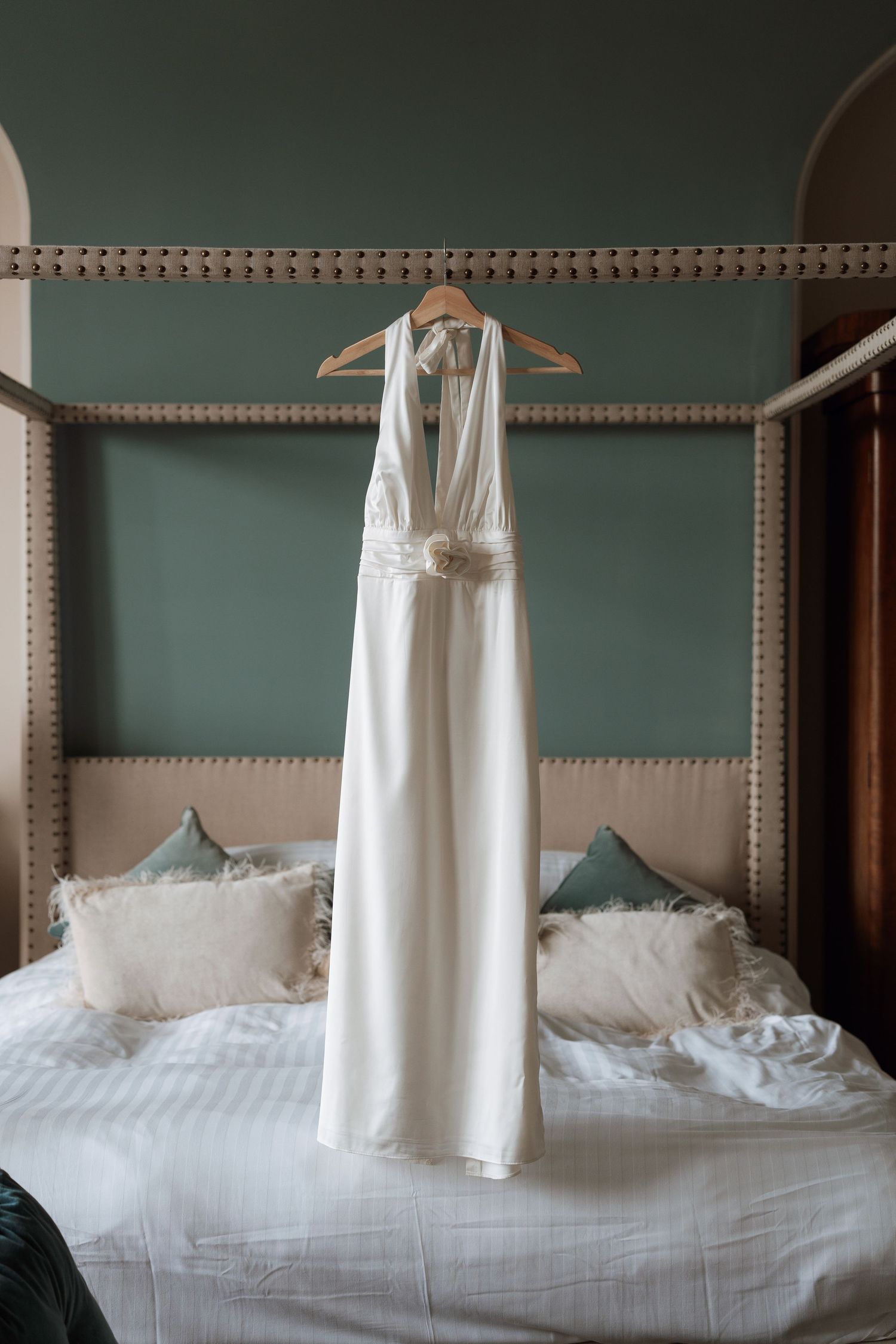 White wedding dress hanging on display against sage green wall in elegant bedroom setting.