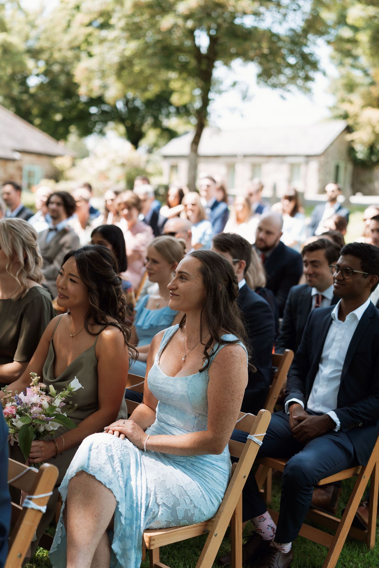 Wedding guests in light blue bridesmaid dresses smile and laugh while seated on wooden chairs during an outdoor ceremony.