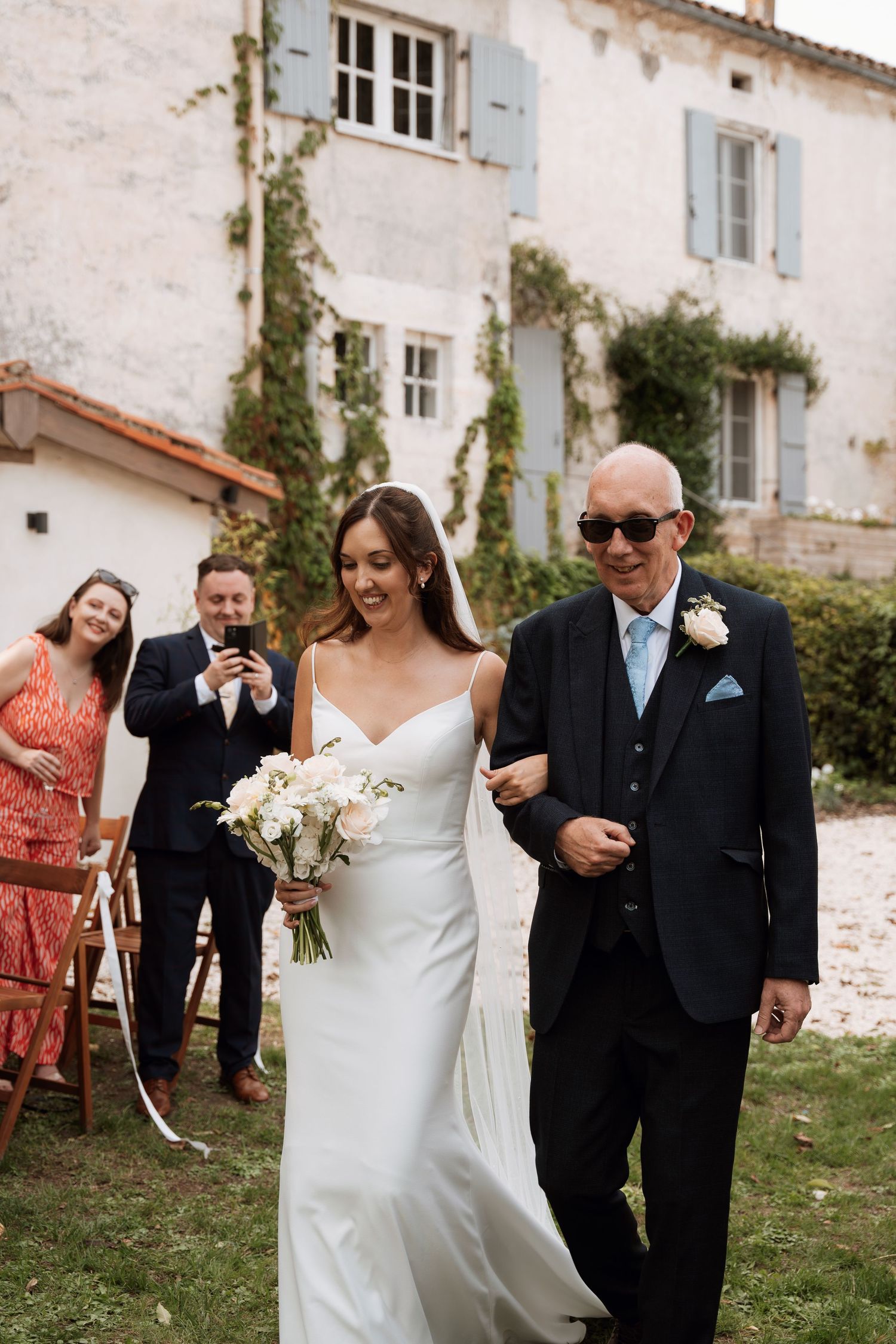 A wedding procession walking along a charming stone building with blue shutters.