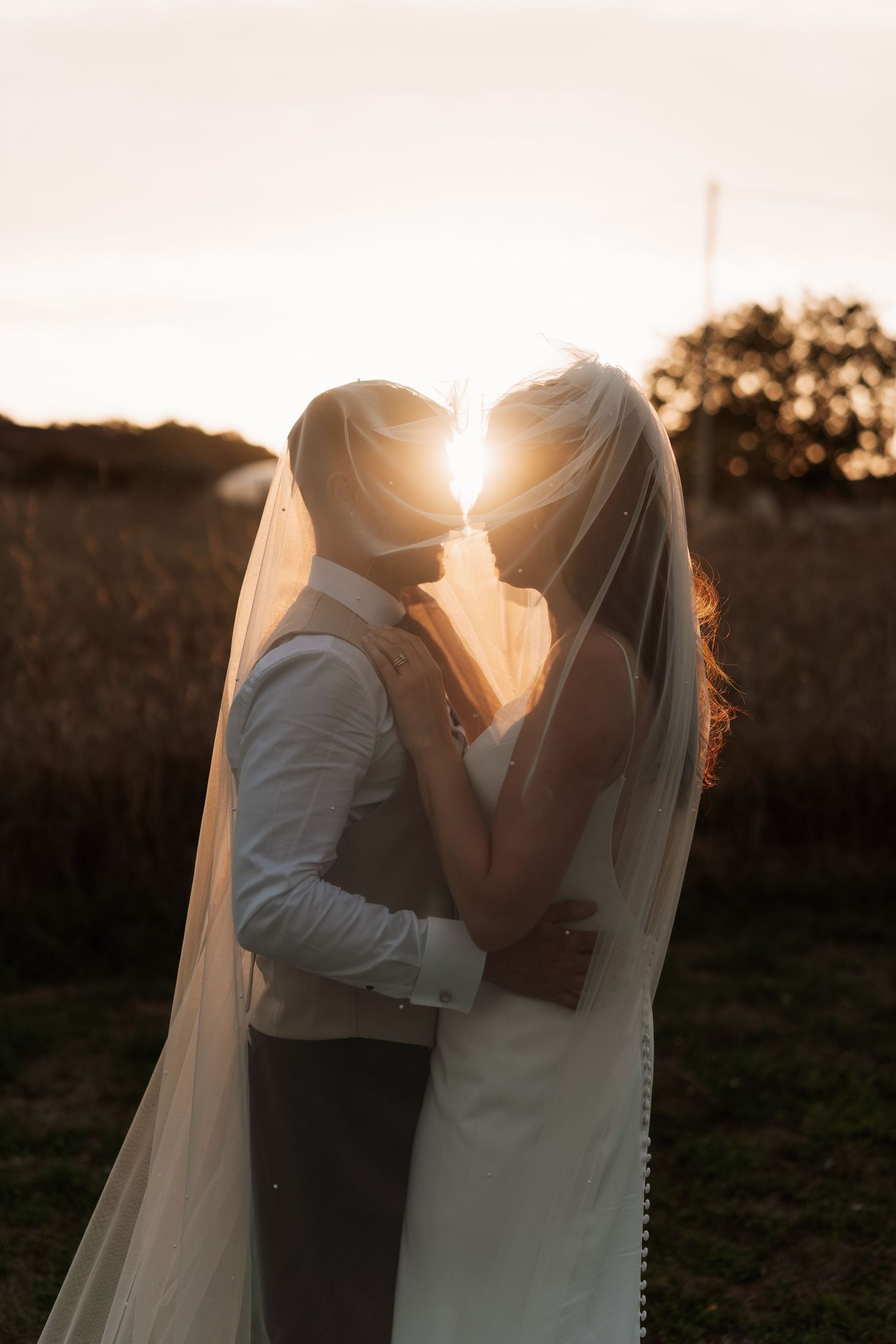 Silhouette of newlyweds kissing at sunset with bride's veil flowing in golden light.