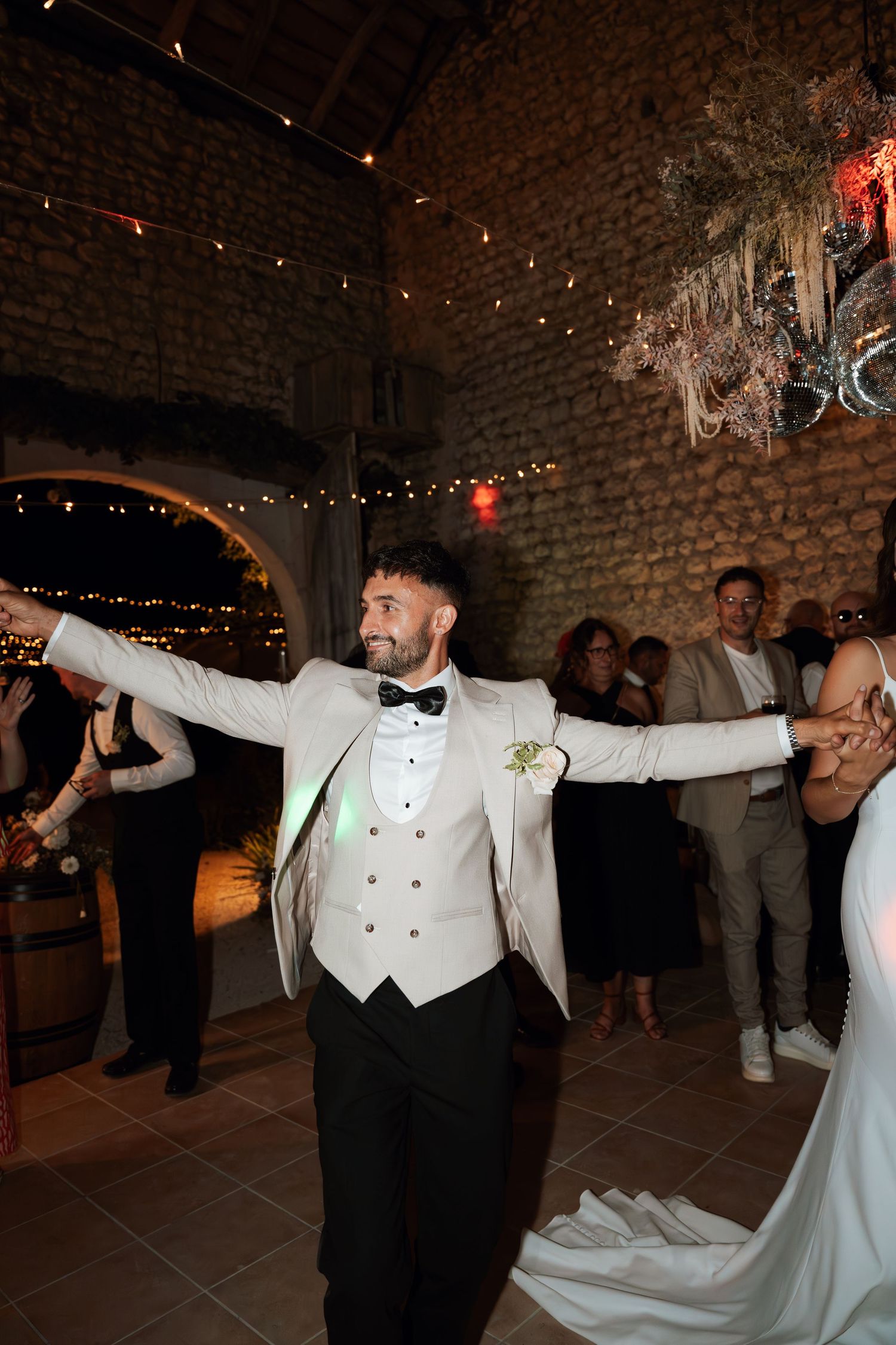 A groom in white tuxedo dances enthusiastically at wedding reception with string lights overhead.