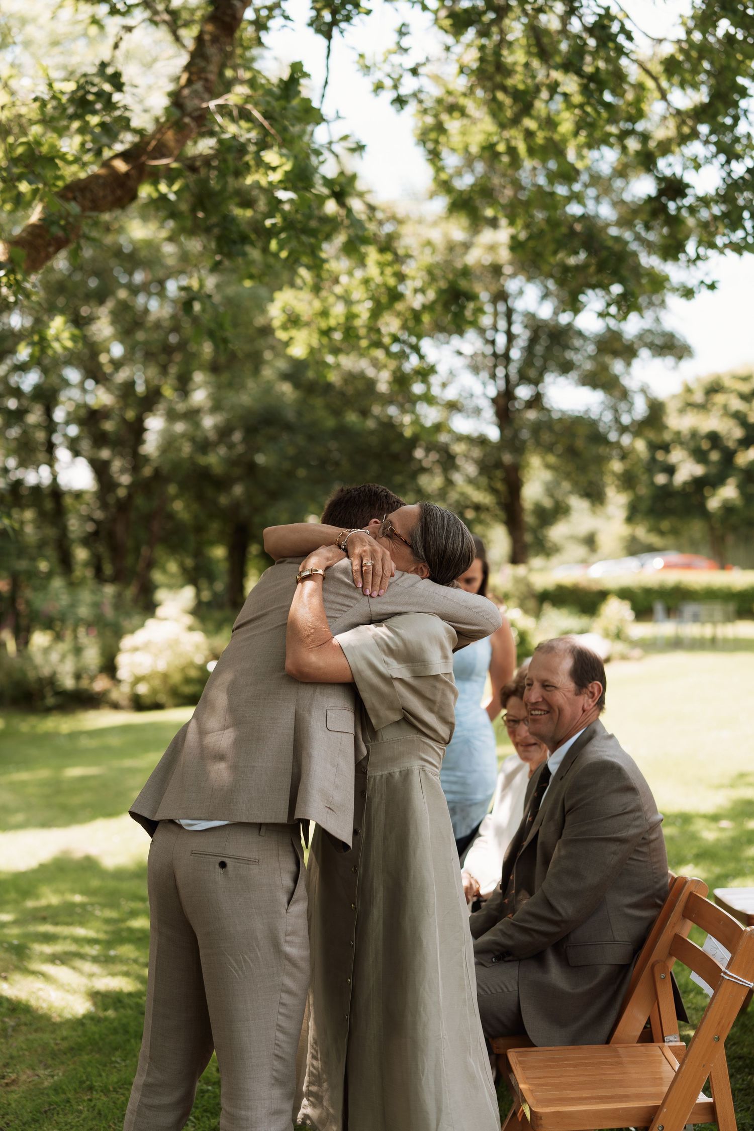 Wedding guests in gray suits embrace during an outdoor celebration.