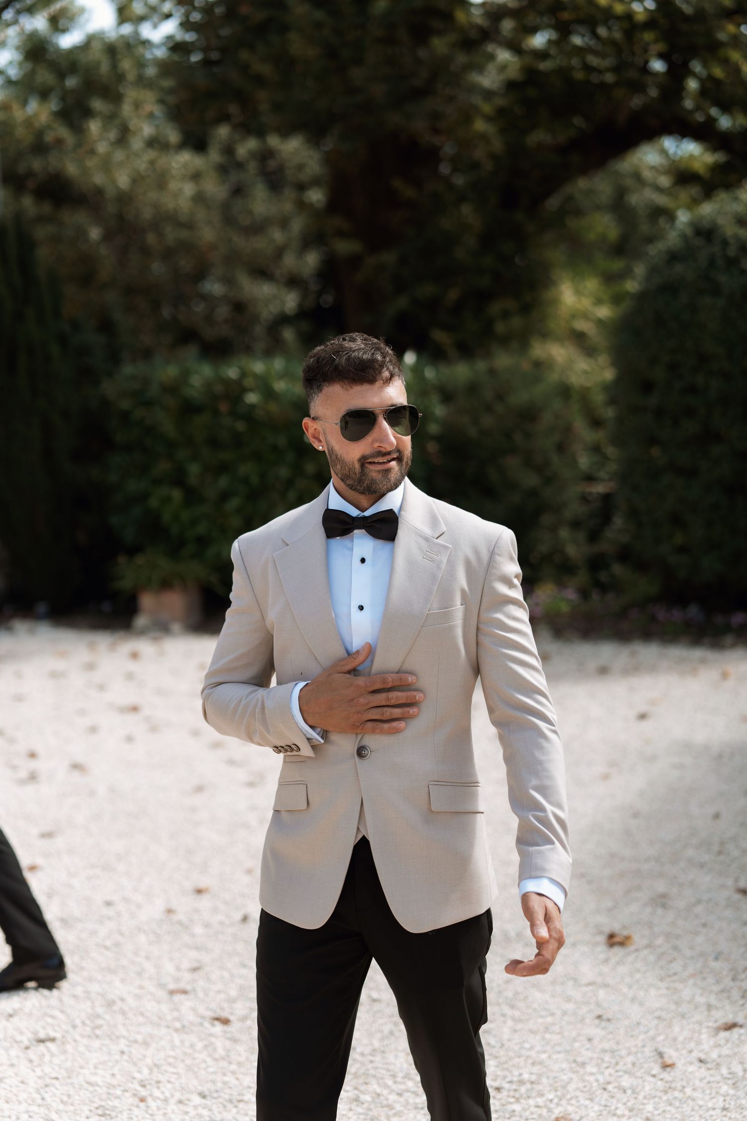 A groom in a light colored tuxedo jacket and bowtie poses outdoors.