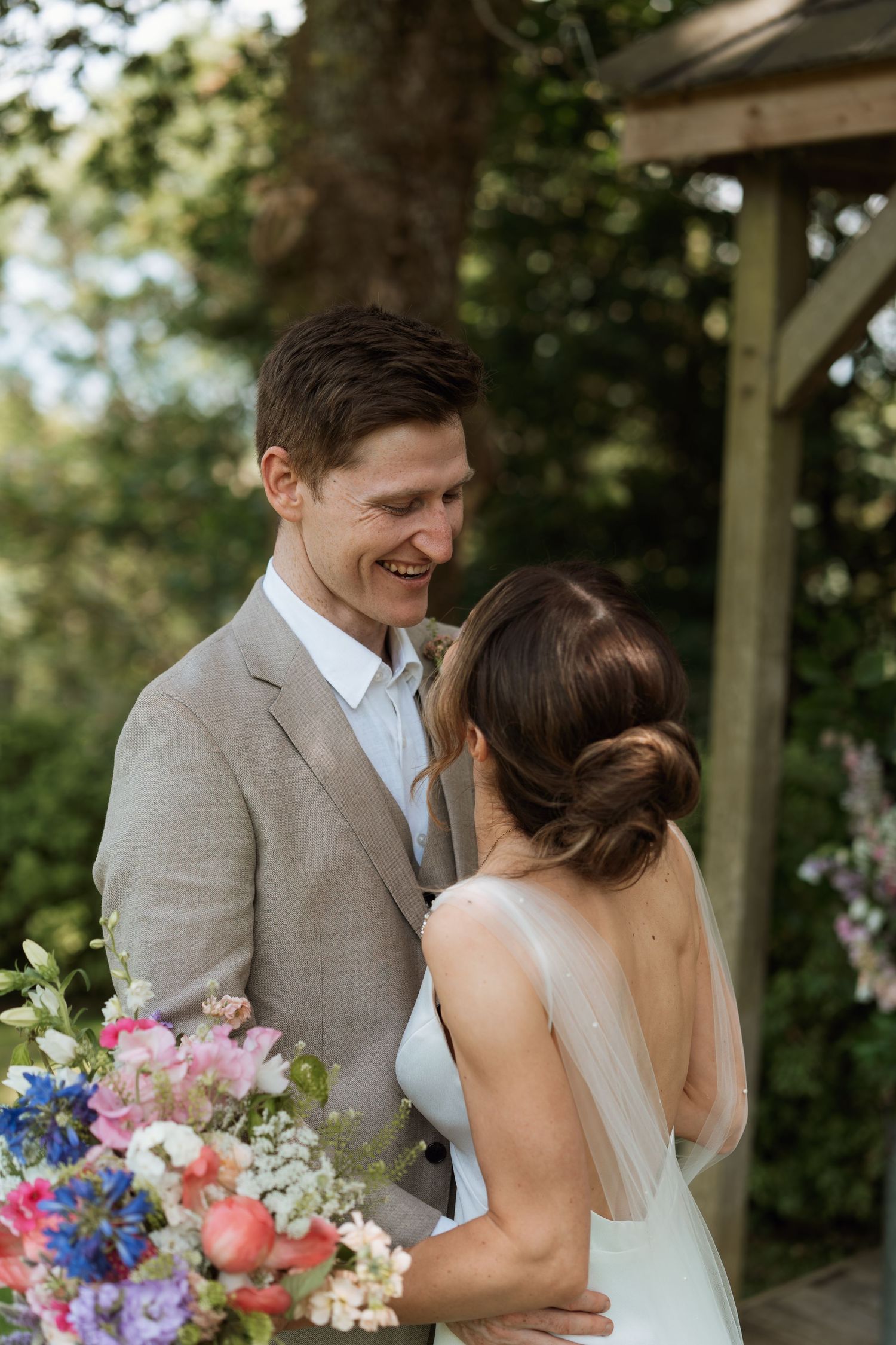 Romantic wedding moment captured under wooden pergola with colorful summer bouquet.